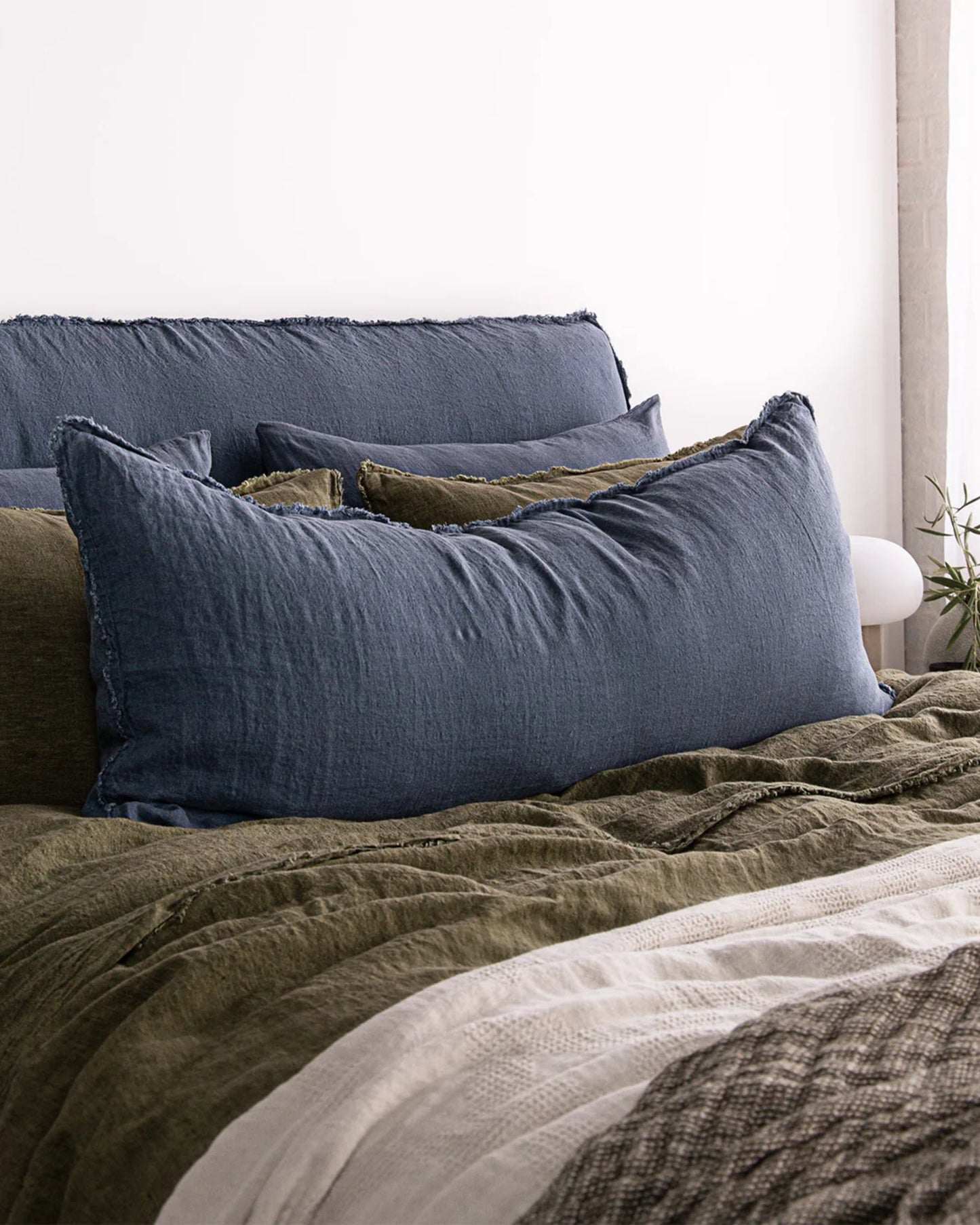 A bed adorned with the Hale Flocca Body Pillow Case from Hale Mercantile Co, featuring a blend of blue and olive green pillows and blankets. The bedding showcases various textures for a cozy, layered look. A white background wall contrasts beautifully with the soft, earthy tones of the bed linens.