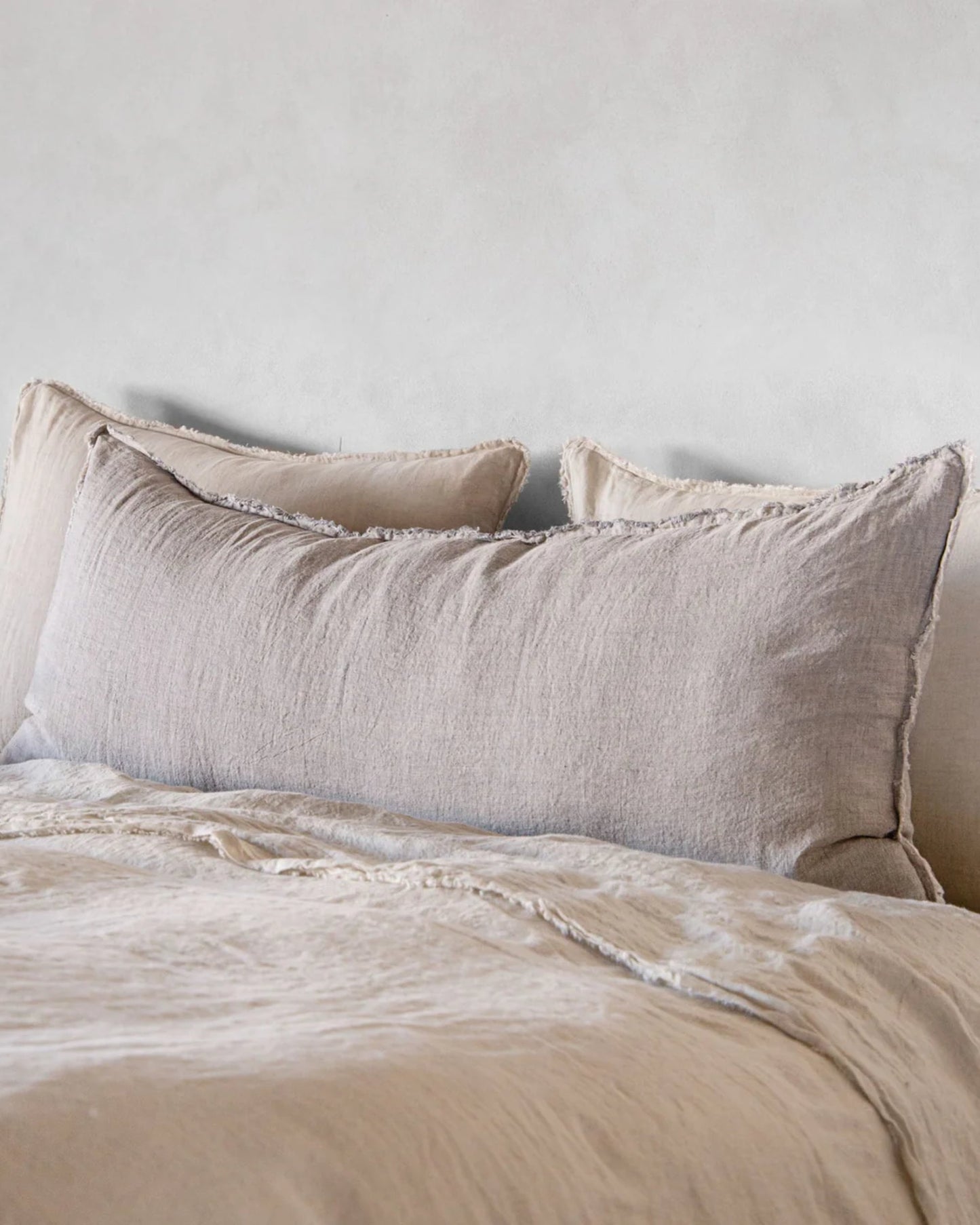 A neatly made bed featuring the Hale Flocca Body Pillow Case by Hale Mercantile Co, with large beige pillows and matching linen bedding against a neutral-colored wall. The pillow cases have a soft, textured appearance, enhancing the cozy and minimalist aesthetic of the room.
