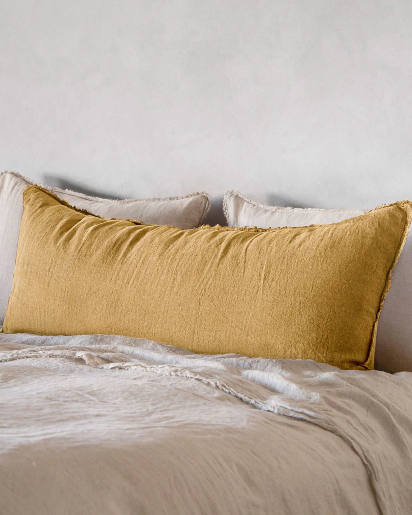 A cozy bedroom scene features the Hale Flocca Body Pillow Case in a large mustard yellow hue resting against neutral-colored pillows on a neatly made bed. The light beige bedding from Hale Mercantile Co creates a soft and inviting atmosphere against a plain white wall.
