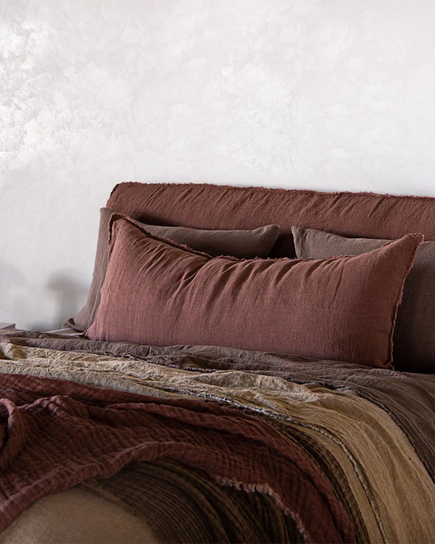 A cozy bed adorned with Hale Mercantile Co's Flocca Body Pillow Case in brown and beige, featuring large pillows in earthy tones. The headboard complements the soft color scheme with simplicity. The ambiance is warm and inviting, set against a textured light gray wall in the background.