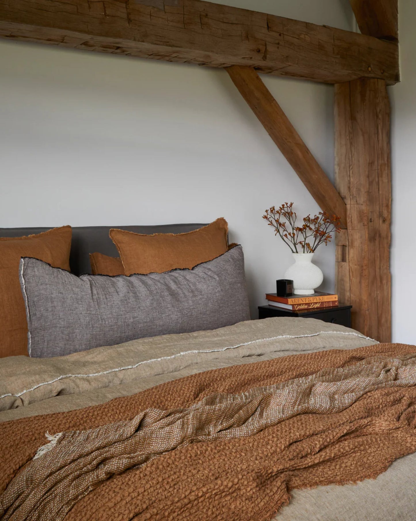 Cozy bedroom interior featuring a wooden beam above the bed. The bed is adorned with pillows covered in Hale Flocca Body Pillow Cases by Hale Mercantile Co, complemented by a beige duvet and a textured brown blanket. A black nightstand holds a white vase filled with dried branches and two stacked books.