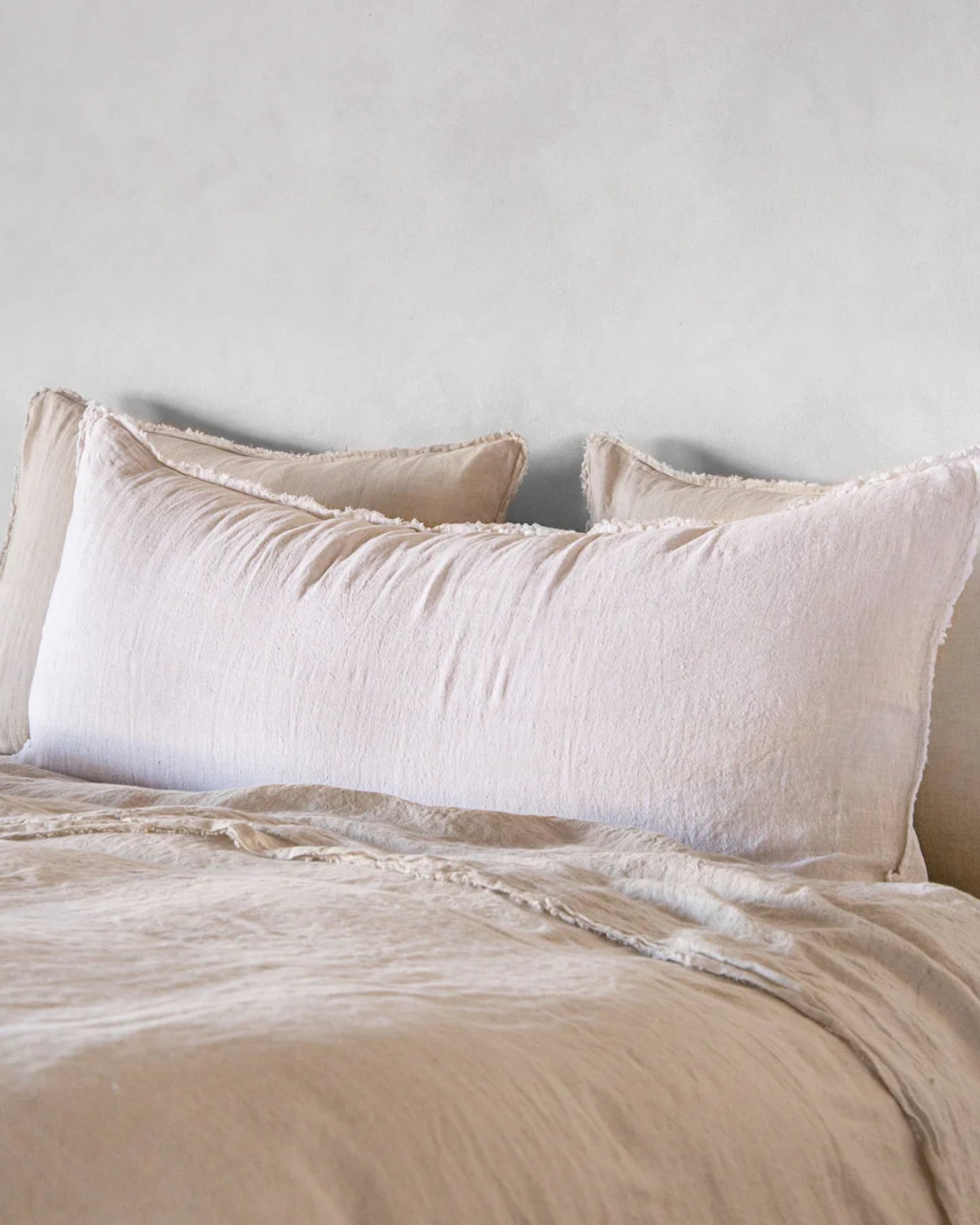 A neatly made bed featuring a large, soft pillow encased in the Hale Mercantile Co's Hale Flocca Body Pillow Case, complemented by a matching comforter against a light gray wall, creating a cozy and minimalist bedroom atmosphere.