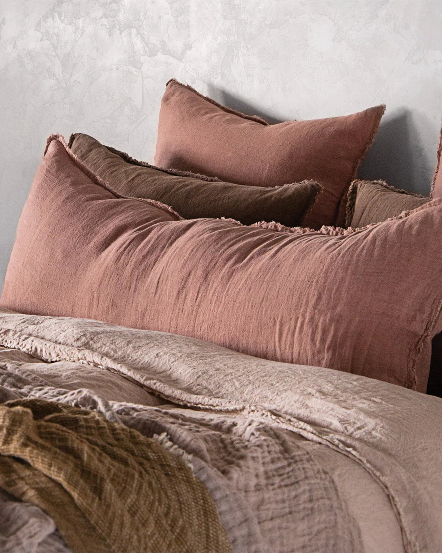 A cozy bedroom setup features a neatly arranged stack of pillows in shades of pink and brown against a light textured wall. The bed is adorned with the Hale Flocca Body Pillow Case by Hale Mercantile Co, complemented by soft pink and beige bedding, creating a warm and inviting atmosphere.