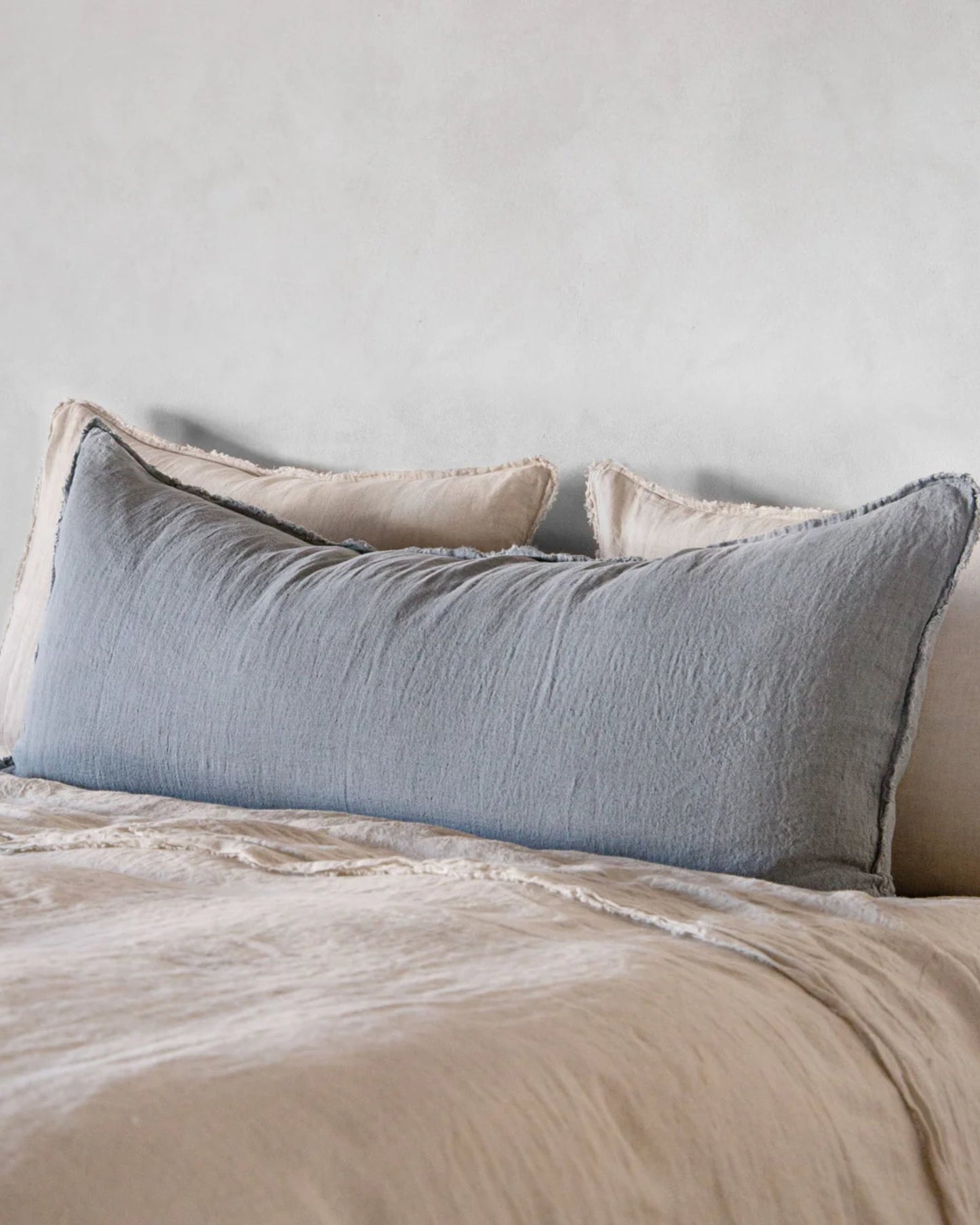 A neatly made bed showcases the Hale Flocca Body Pillow Case in light blue from Hale Mercantile Co, set against a collection of cream-colored pillows and a matching bedspread, all against a backdrop of a light gray wall. The bedding creates a serene and inviting atmosphere.