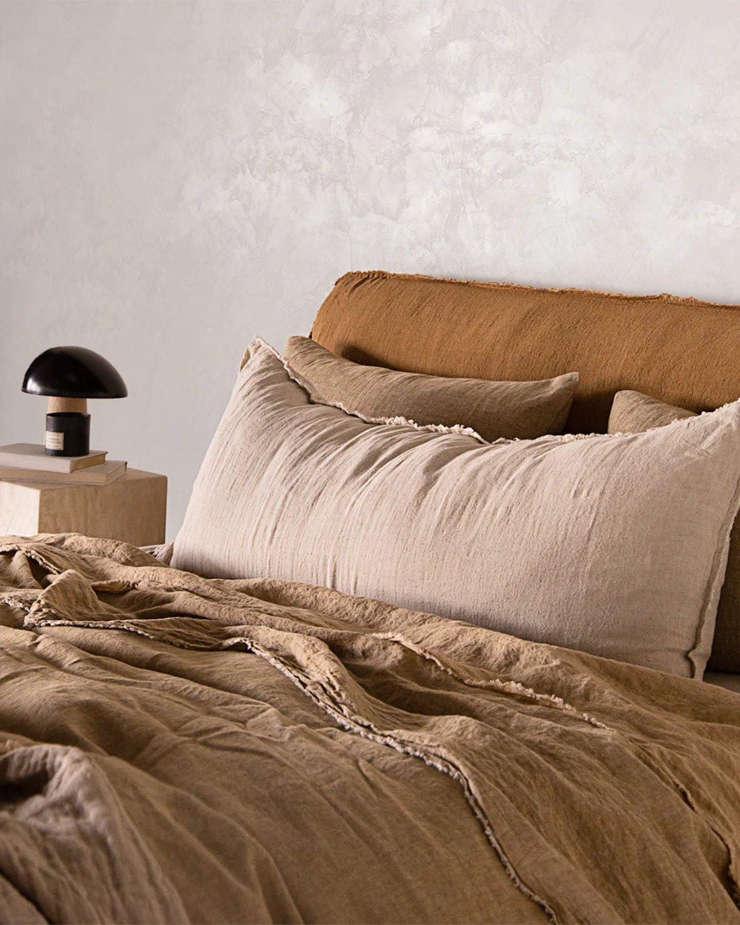 A cozy bedroom showcases a bed adorned with beige linen sheets and the Hale Flocca Body Pillow Case from Hale Mercantile Co. A small black and white lamp rests on a wooden nightstand, while the light-colored textured wall infuses the space with a warm and serene ambiance.