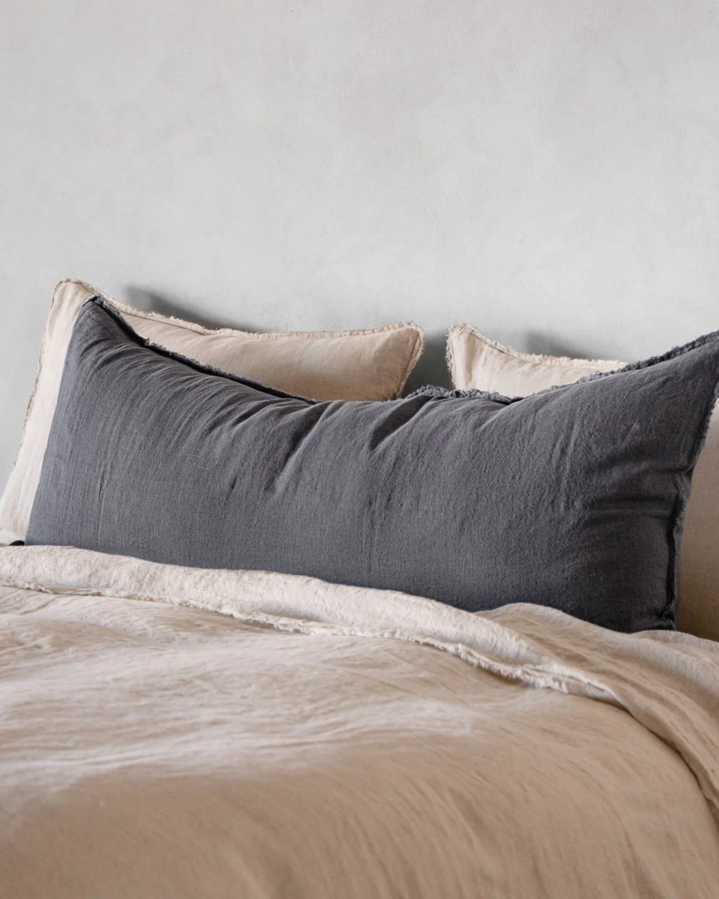 A neatly made bed featuring cream-colored sheets and pillows is complemented by the Hale Flocca Body Pillow Case in dark gray from Hale Mercantile Co. The large bolster pillow takes center stage in front of two lighter pillows, set against a light gray wall, creating a cozy and minimalist bedroom setup.