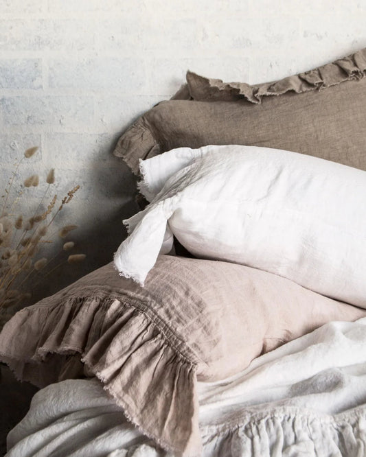 A cozy bed setup highlights a stack of HALE KRISTINE EURO PILLOWCASES by Hale Mercantile Co, featuring earthy tones with ruffled edges, perfectly paired with an easy-care linen bedding set. The light-colored, textured wall and dried flowers enhance the vintage-inspired bedroom decor's rustic ambiance.