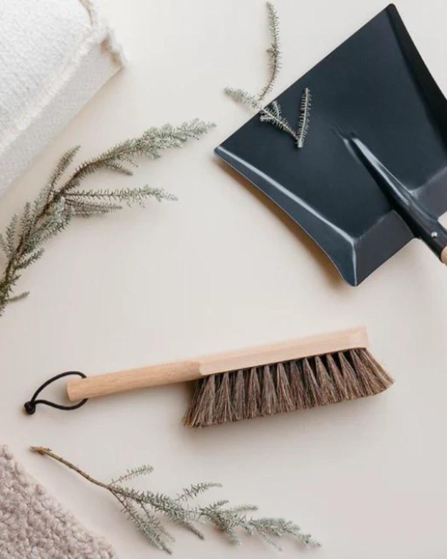 A HAND BRUSH by Andree Jardin, featuring a wooden handle and natural bristles, rests on a clean surface, surrounded by sprigs of greenery. Nearby, textured beige fabrics enhance the handcrafted, minimalist aesthetic.