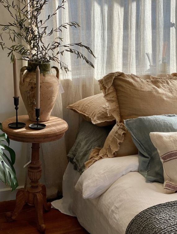 A cozy bedroom corner featuring a bed with white and beige pillows, a wooden side table with two tall candlesticks, and a large vase holding branches. Sheer curtains allow light to filter in, creating a warm, inviting atmosphere.