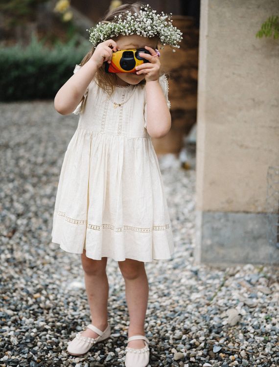 A young girl in a cream dress and floral crown stands on a gravel path, holding a toy camera to her eye. She appears to be taking a photo. The background features a textured wall and some greenery.