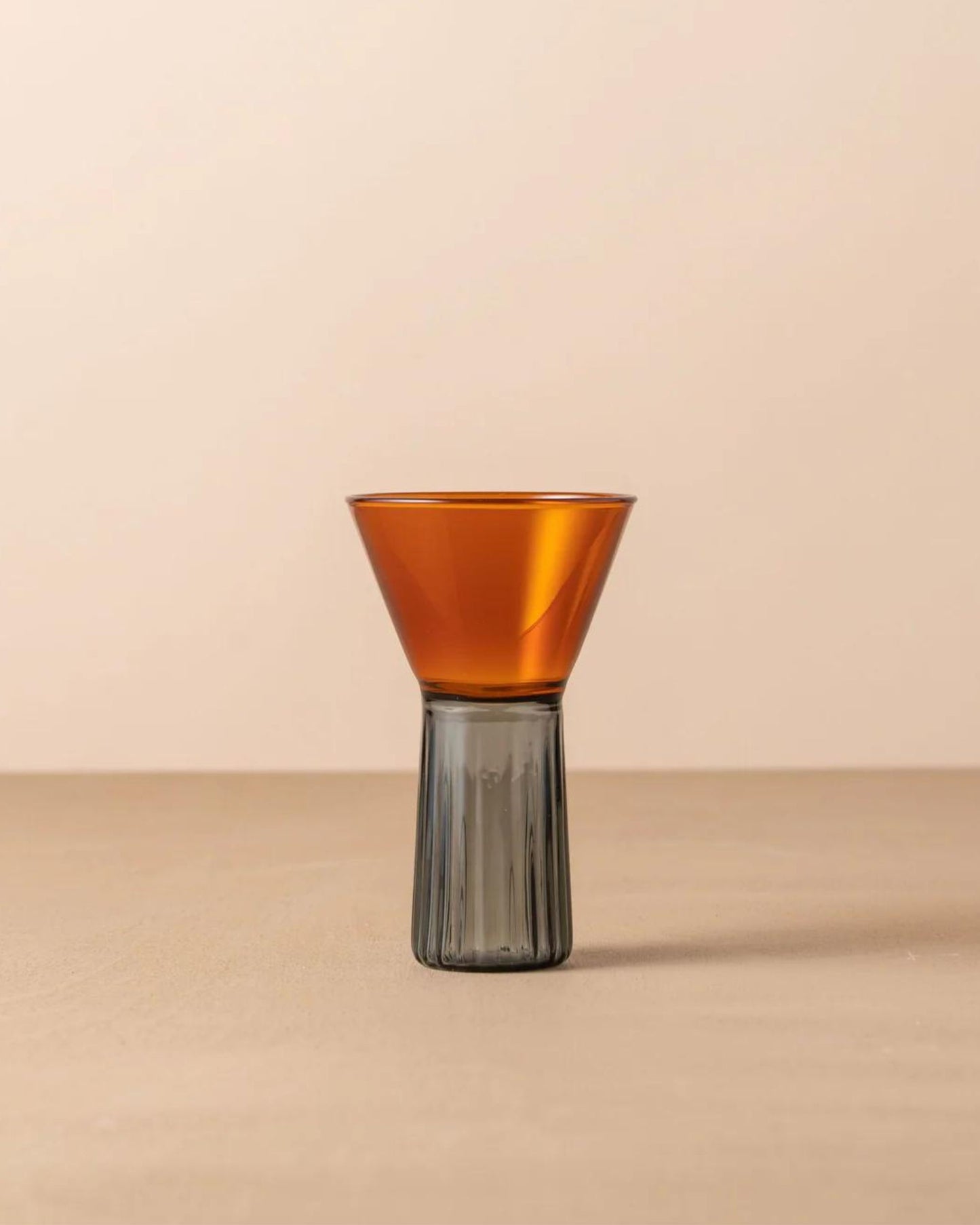 The KAIROS WINE GLASS by Saarde, featuring a small, modern design with a brown cone-shaped top and a gray ribbed cylindrical base inspired by vintage Murano glassware, is displayed on a beige surface against a neutral background.