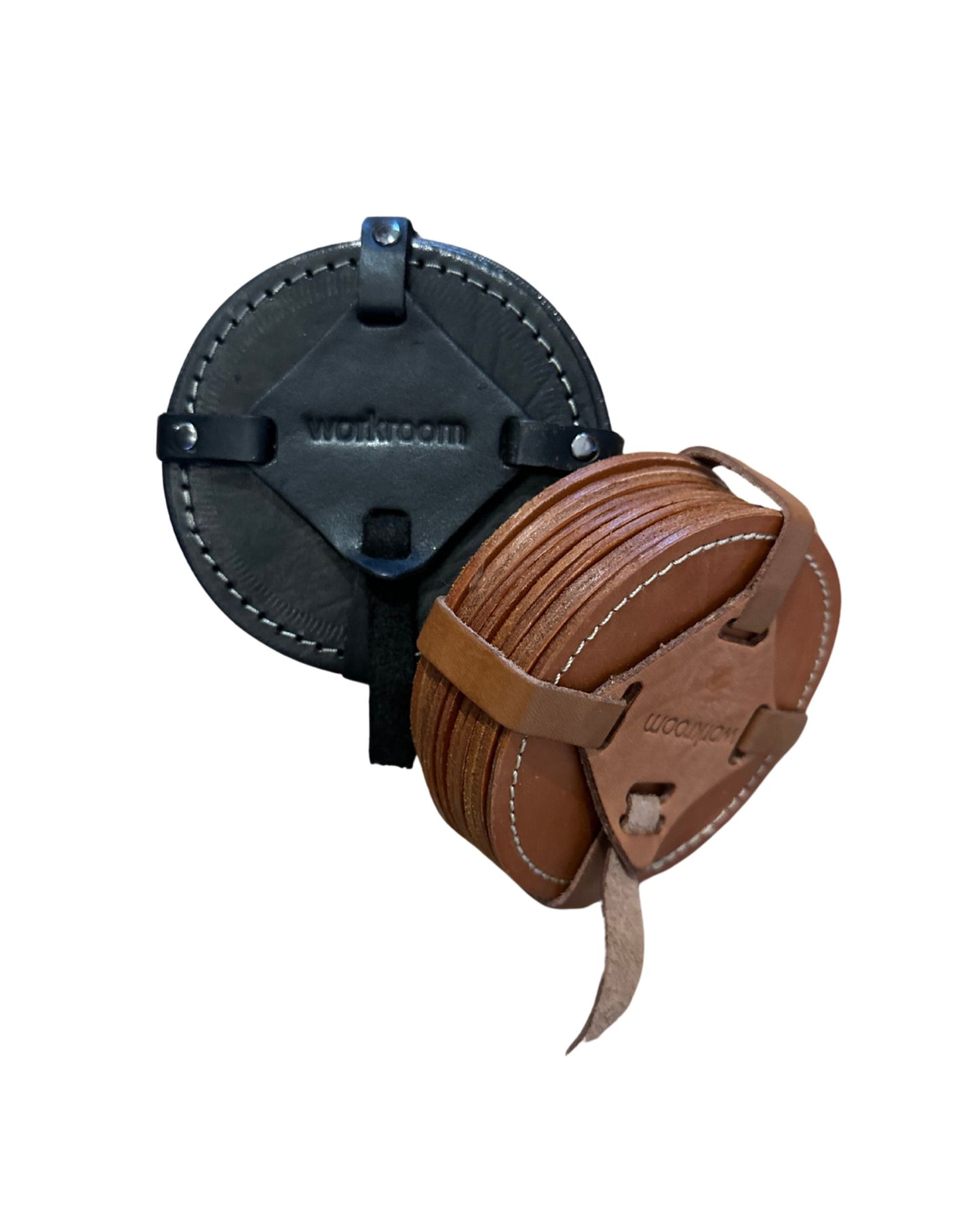 A set of WORKROOM LEATHER COASTERS from The Workroom, featuring a minimalist design with visible stitching for added elegance and durability. Each stack, secured with straps, includes one set in black full-grain leather and another in brown, embossed with "workroom" on every coaster.