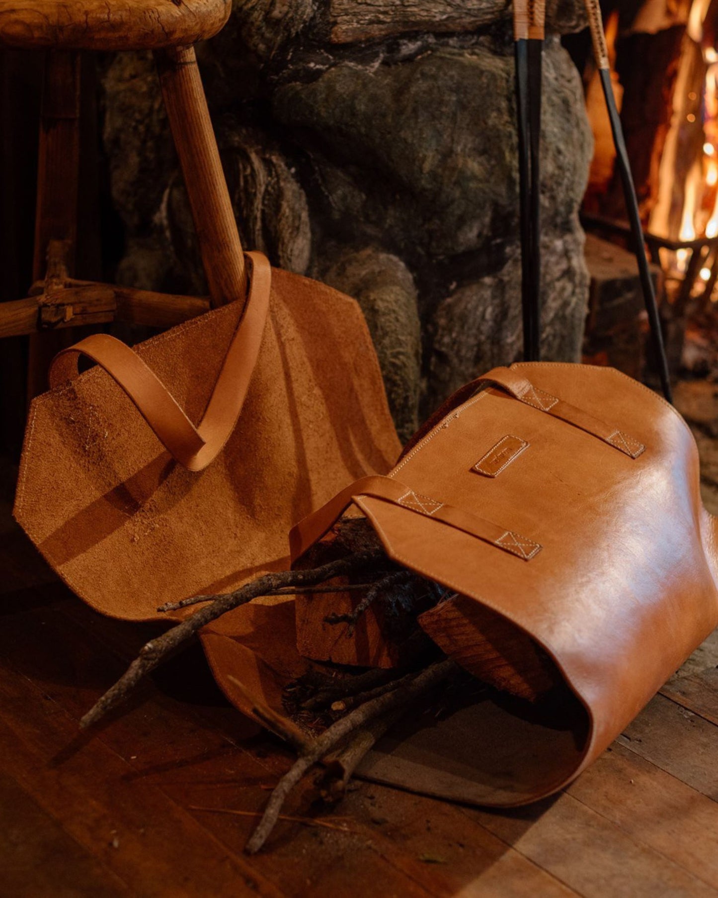 A Leather Log Carrier by The Workroom, filled with firewood, rests against a rustic stone fireplace. A wooden stool is nearby, and a fire glows warmly in the background, creating a cozy atmosphere.