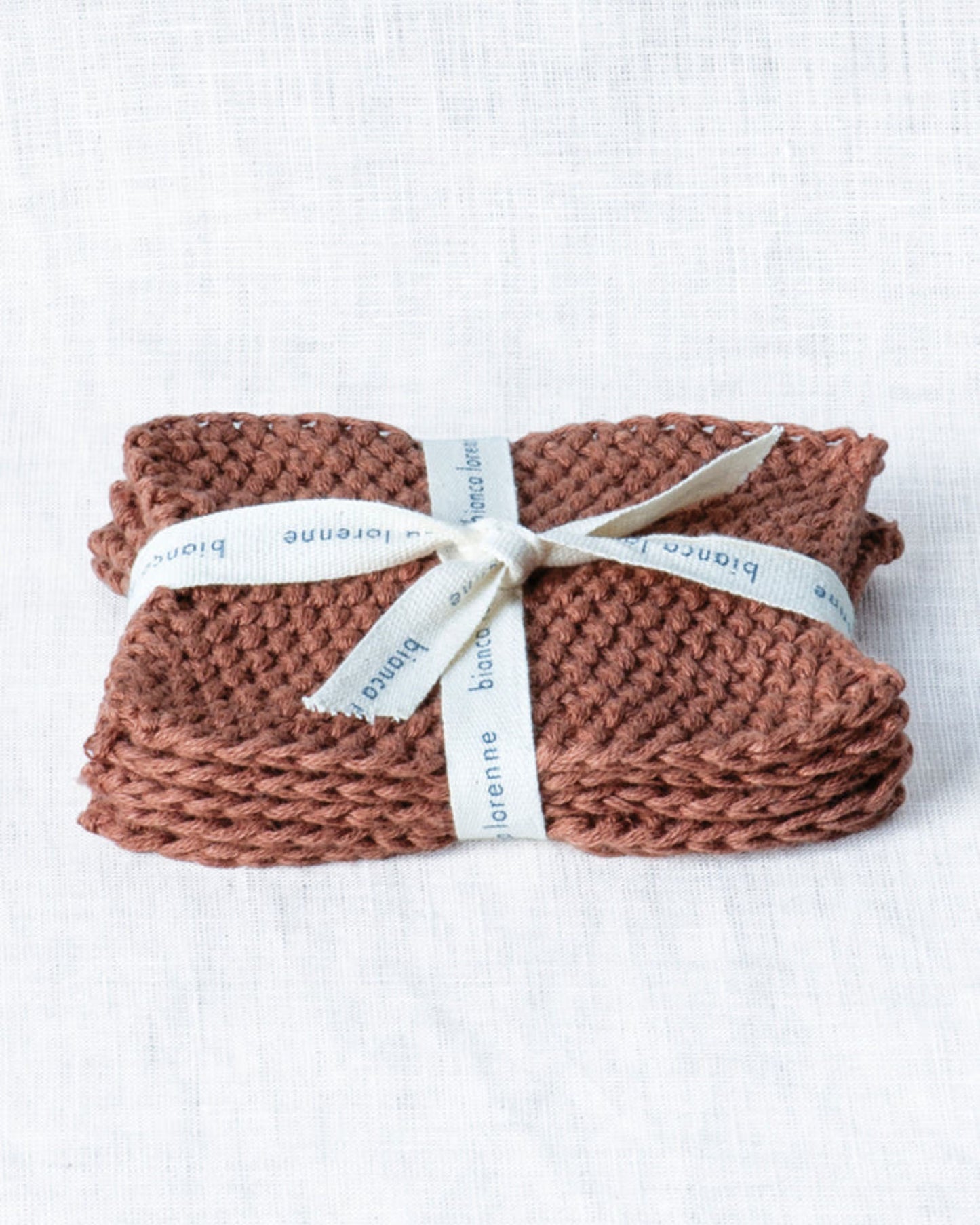 A brown, knitted washcloth crafted from organic cotton is neatly folded and tied with an off-white ribbon embossed with the brand name Bianca Lorenne. This sustainable choice, ideal as a reusable makeup remover called Lavette Makeup Remover, rests on a textured white surface.