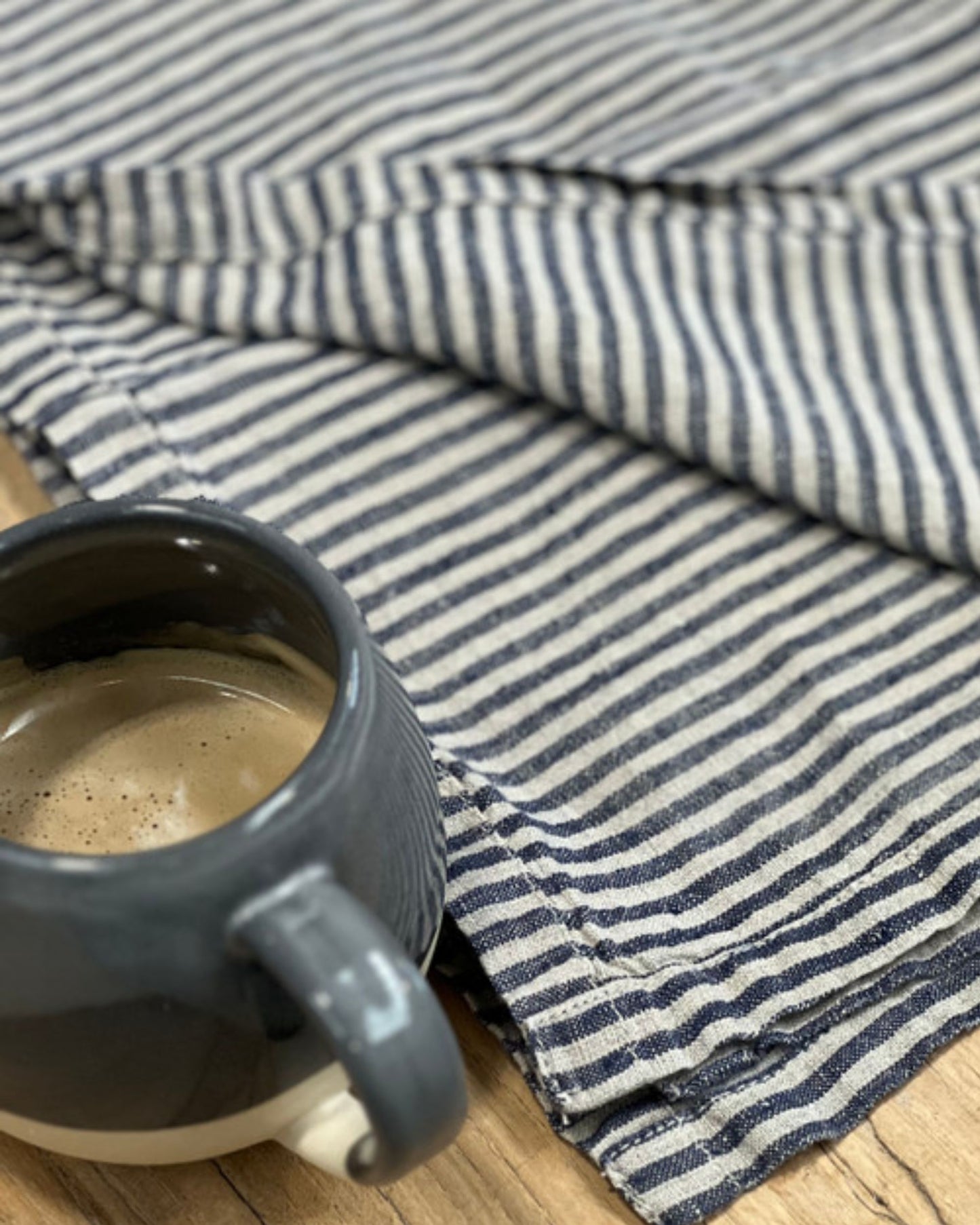 A dark ceramic mug filled with coffee sits on a wooden table next to a folded NOEMI BLUE STRIPE TEA TOWEL by Rustic Linen, adding a touch of cozy elegance to your kitchen essentials.