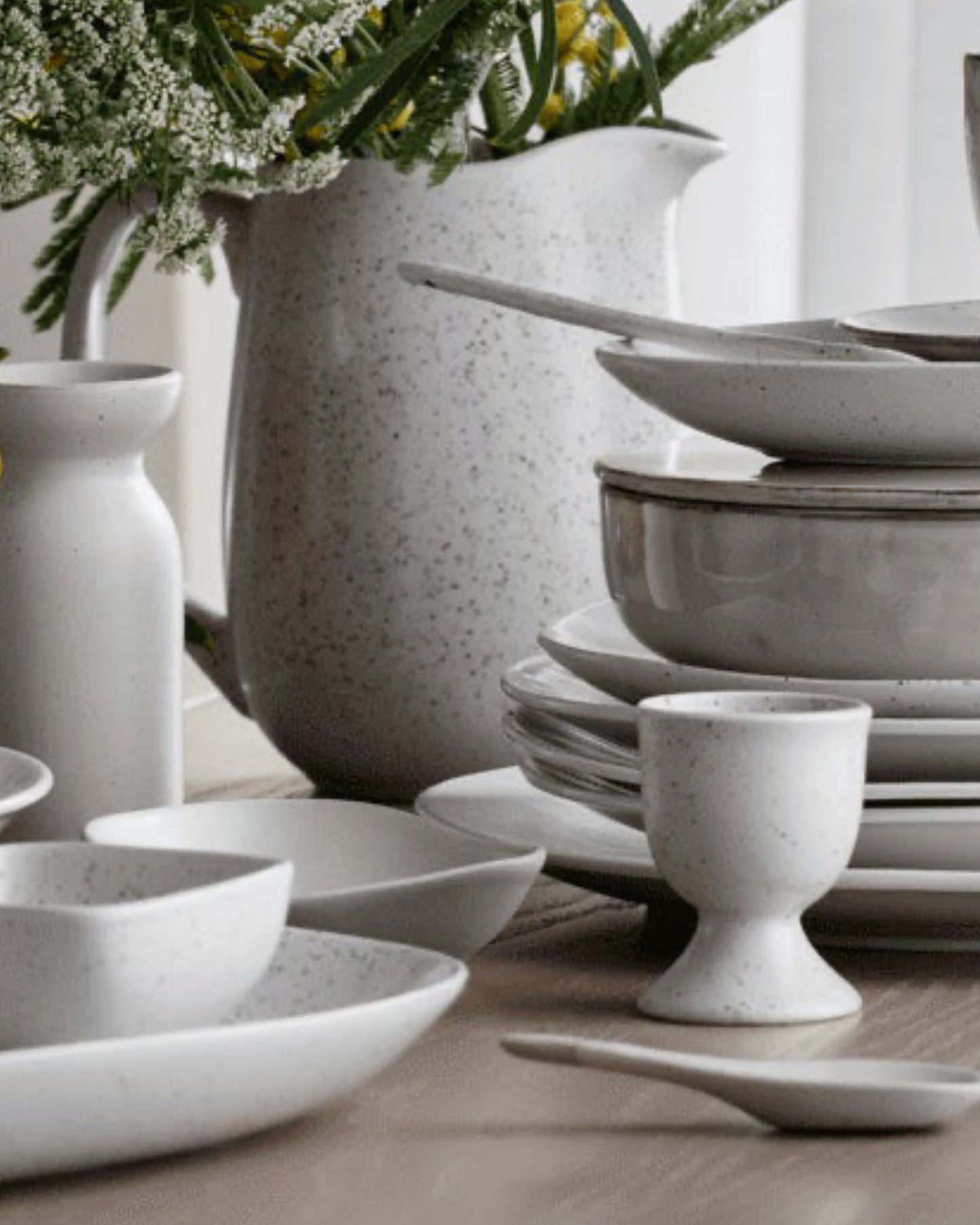 A Broste EGG CUP, along with rustic white ceramic plates, bowls, and cups, is artfully displayed on a wooden surface. The speckled design of the dishes pairs harmoniously with flowers in the background, capturing the allure of a cozy breakfast setting.