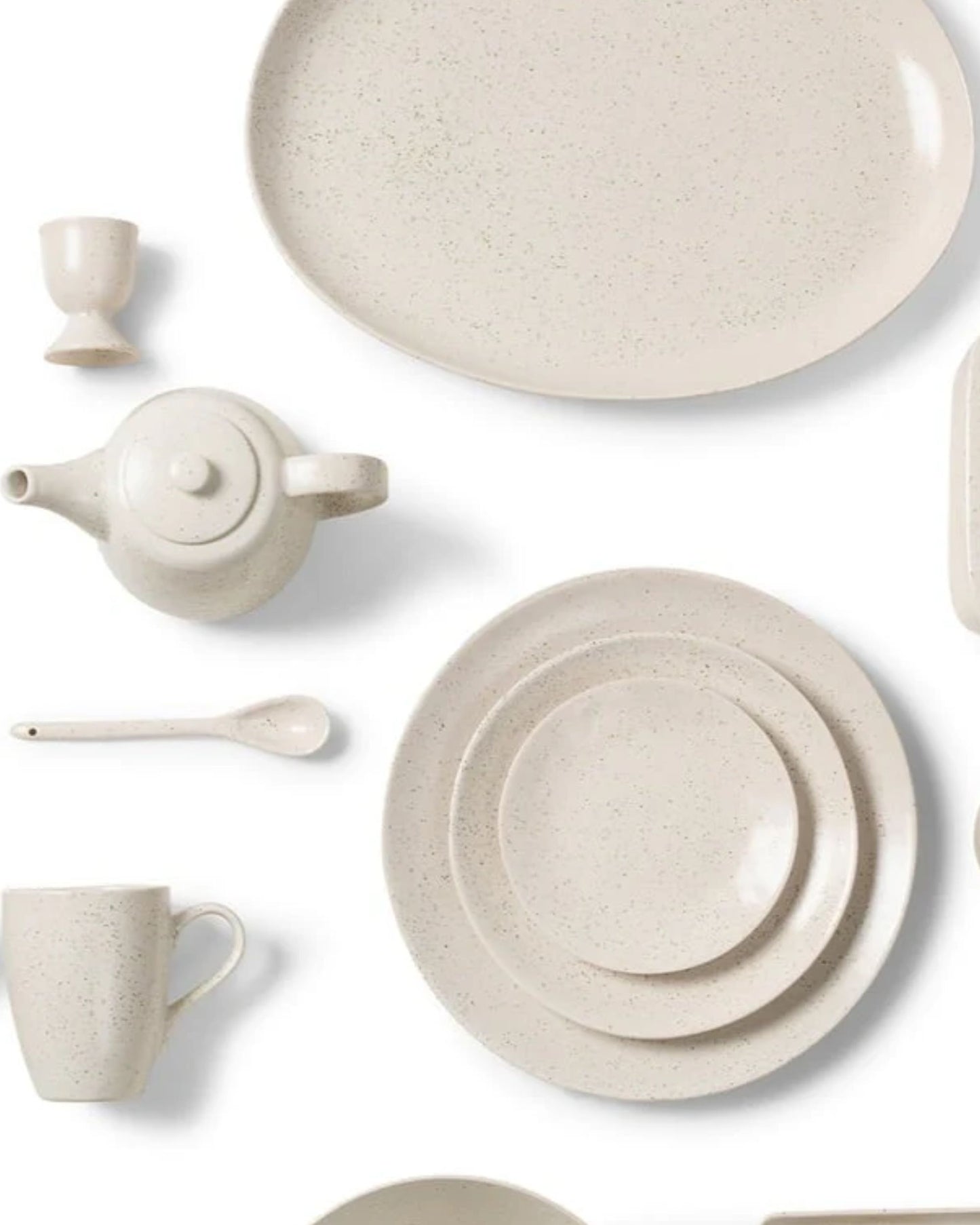 A beautifully arranged collection of Broste's speckled beige ceramic dishware, featuring a teapot, plates, a cup, a spoon, an EGG CUP, and an oval serving platter, graces the breakfast table against a crisp white background.