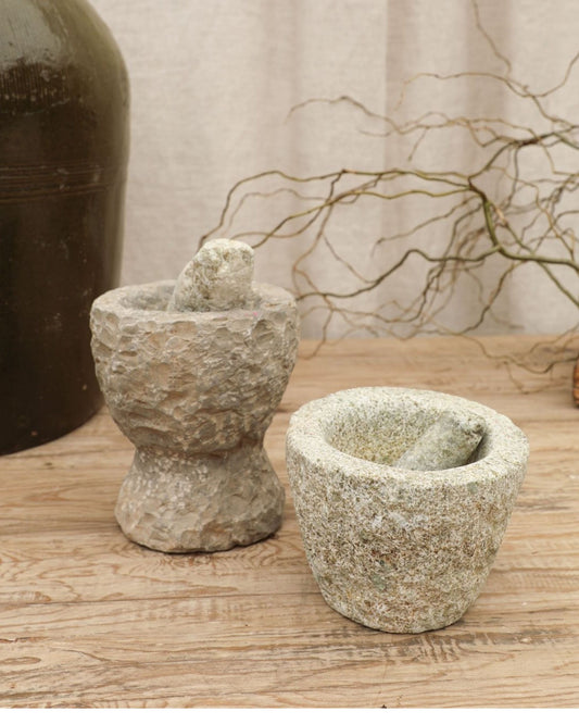 Two Original Stone Mortars by Hawthorne are placed on a wooden surface. The taller mortar on the left features a textured finish, while the shorter one has a smoother surface. In the background, a large ceramic pot and dried branches enhance the earthy ambiance.
