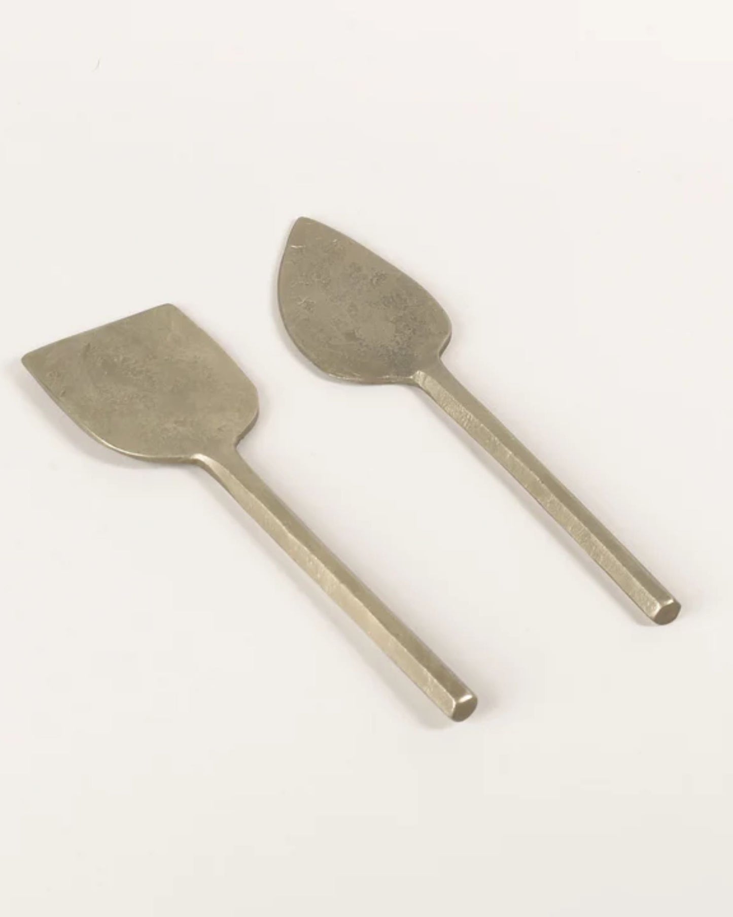 Two silver antique cheese knives from the New Direction brand, collectively named the PARM KNIFE, are unique handmade items with distinct blade shapes. One knife features a broad, flat blade while the other has a narrow, teardrop-shaped blade. Both boast simple, straight handles that are lovingly preserved with food-safe oil to ensure enduring elegance and functionality.