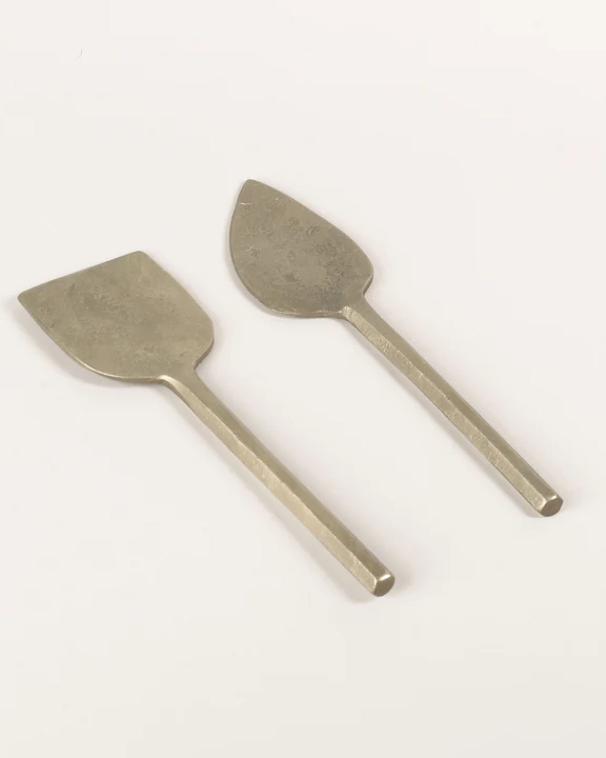 Two silver antique cheese knives from the New Direction brand, collectively named the PARM KNIFE, are unique handmade items with distinct blade shapes. One knife features a broad, flat blade while the other has a narrow, teardrop-shaped blade. Both boast simple, straight handles that are lovingly preserved with food-safe oil to ensure enduring elegance and functionality.