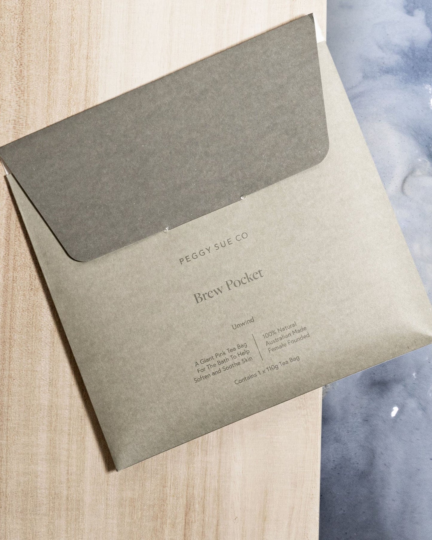 A PEGGY SUE - BREW POCKET by Peggy Sue rests on a wooden surface. This reusable, 100% natural tea bag enriches self-care routines with its refillable design. The minimalist packaging casts soft shadows, elegantly highlighting the natural ingredients against a textured background.