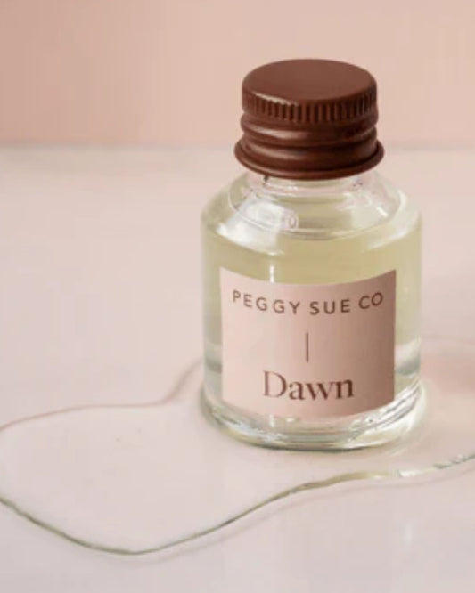 A small, clear glass bottle labeled PEGGY SUE - ESSENTIAL OIL PERFUME by Peggy Sue with a brown cap rests on a light surface. A light-colored liquid, suggesting an uplifting blend of essential oil perfumes, is spilled around it. The background features a soft peach color, enhancing the scene's serene aura.