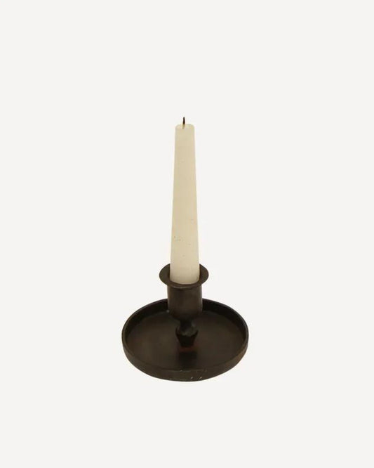 A white taper candle is elegantly set within a RAINE TAPER BLACK CANDLE CUP by French Country, featuring a dark, hand-forged metal design with a broad base reminiscent of an elegant candle pan, all placed against a plain light-colored background.