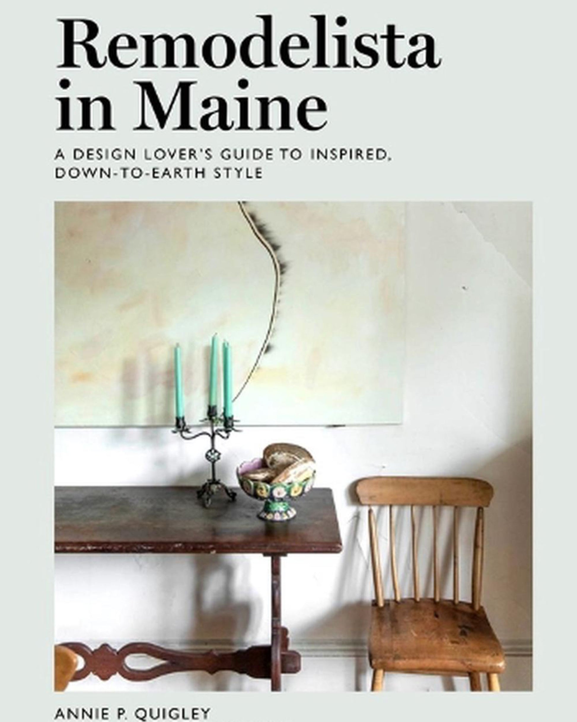 The book cover of "REMODELISTA IN MAINE" from Nation Wide Books, designed by Annie P. Quigley, features a wooden table with a bowl of fruit, green candles, and a wooden chair, capturing the essence of Maine. The white wall decorated with abstract art makes it an ideal choice for design-minded travelers looking for Maine-inspired home decor.