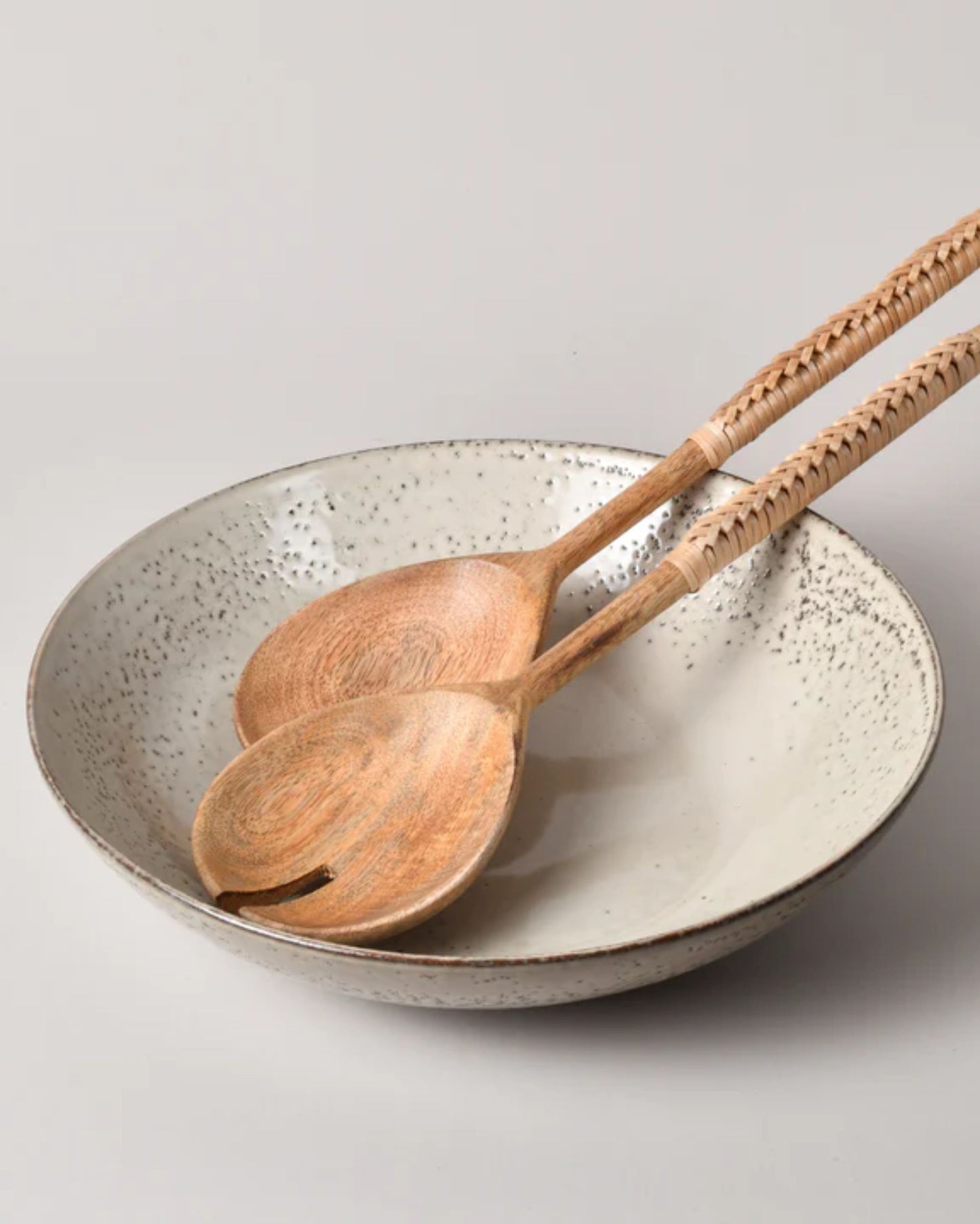 A pair of "SALAD SERVER W/RATTAN HANDLE" by New Direction rests on a speckled ceramic bowl, which exudes a natural, earthy appearance against a plain background.