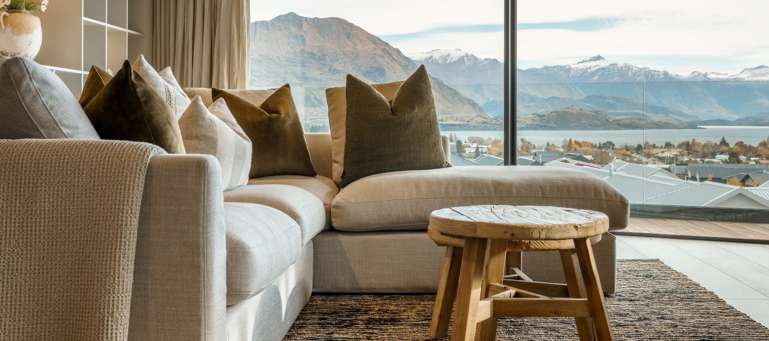 A cozy living room with a beige sectional sofa adorned with green pillows, a round wooden coffee table, and a large window showcasing a scenic view of mountains and a lake. Shelves with decor items are on the left.