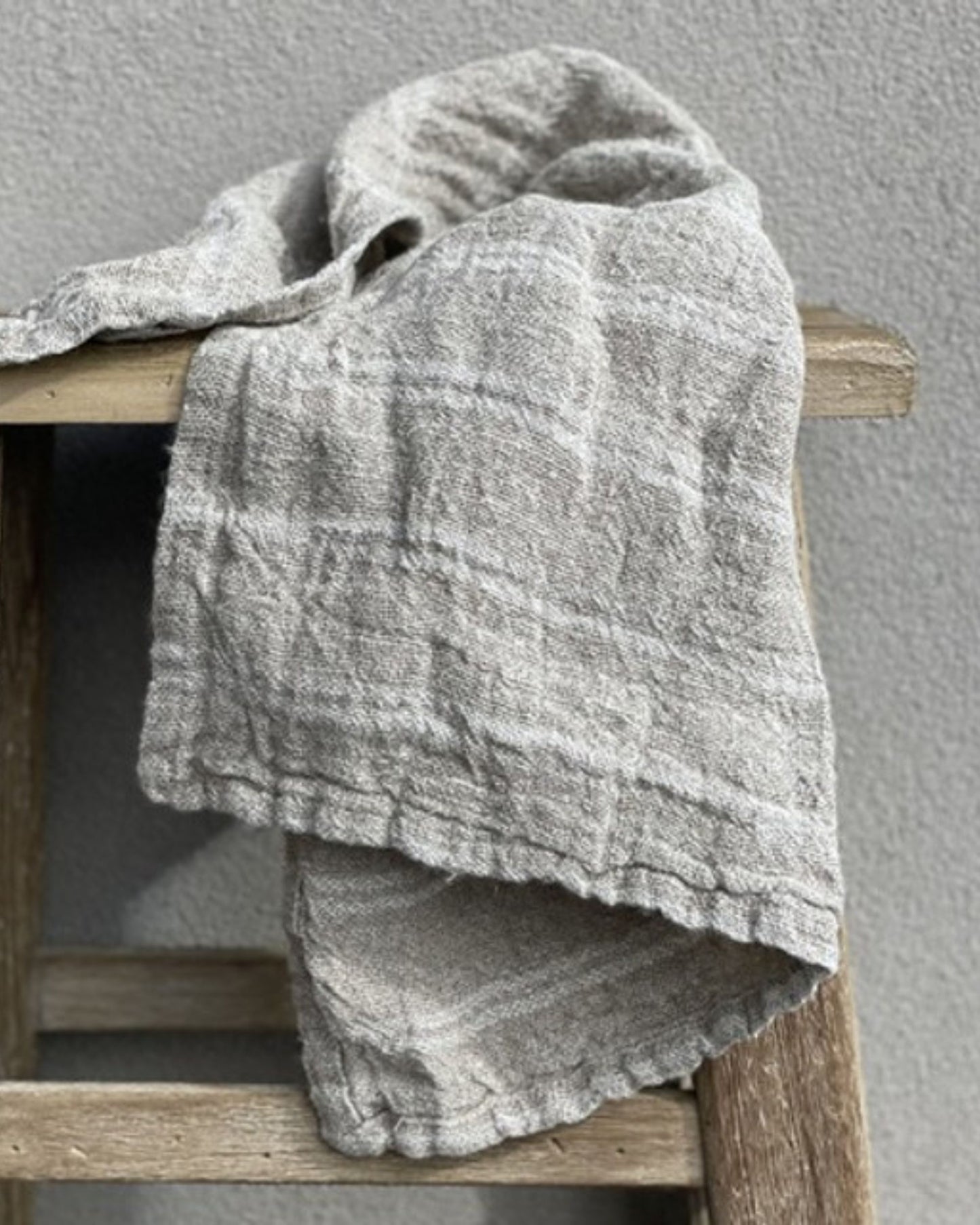 A handloomed tea towel with white stripes from Rustic Linen is elegantly draped over a wooden chair set against a plain gray wall. The fabric's understated pattern exudes a soft, casual charm that adds an element of rustic elegance to the setting.
