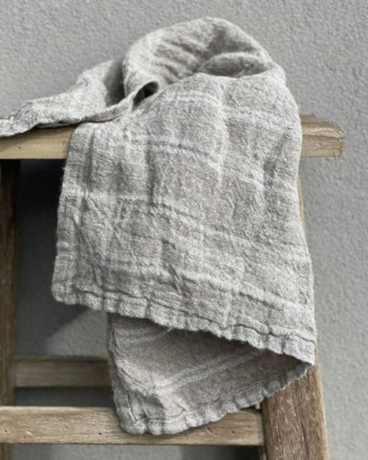 A handloomed tea towel with white stripes from Rustic Linen is elegantly draped over a wooden chair set against a plain gray wall. The fabric's understated pattern exudes a soft, casual charm that adds an element of rustic elegance to the setting.