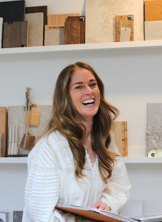 A woman with long brown hair, wearing a white blouse, is smiling and holding a folder. She is standing in front of shelves displaying various tile and wood samples.