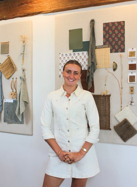 A person in a white dress stands smiling in front of a display board with various textile samples and materials pinned to it. The display includes swatches of fabric, ropes, and design concepts on a neutral background.