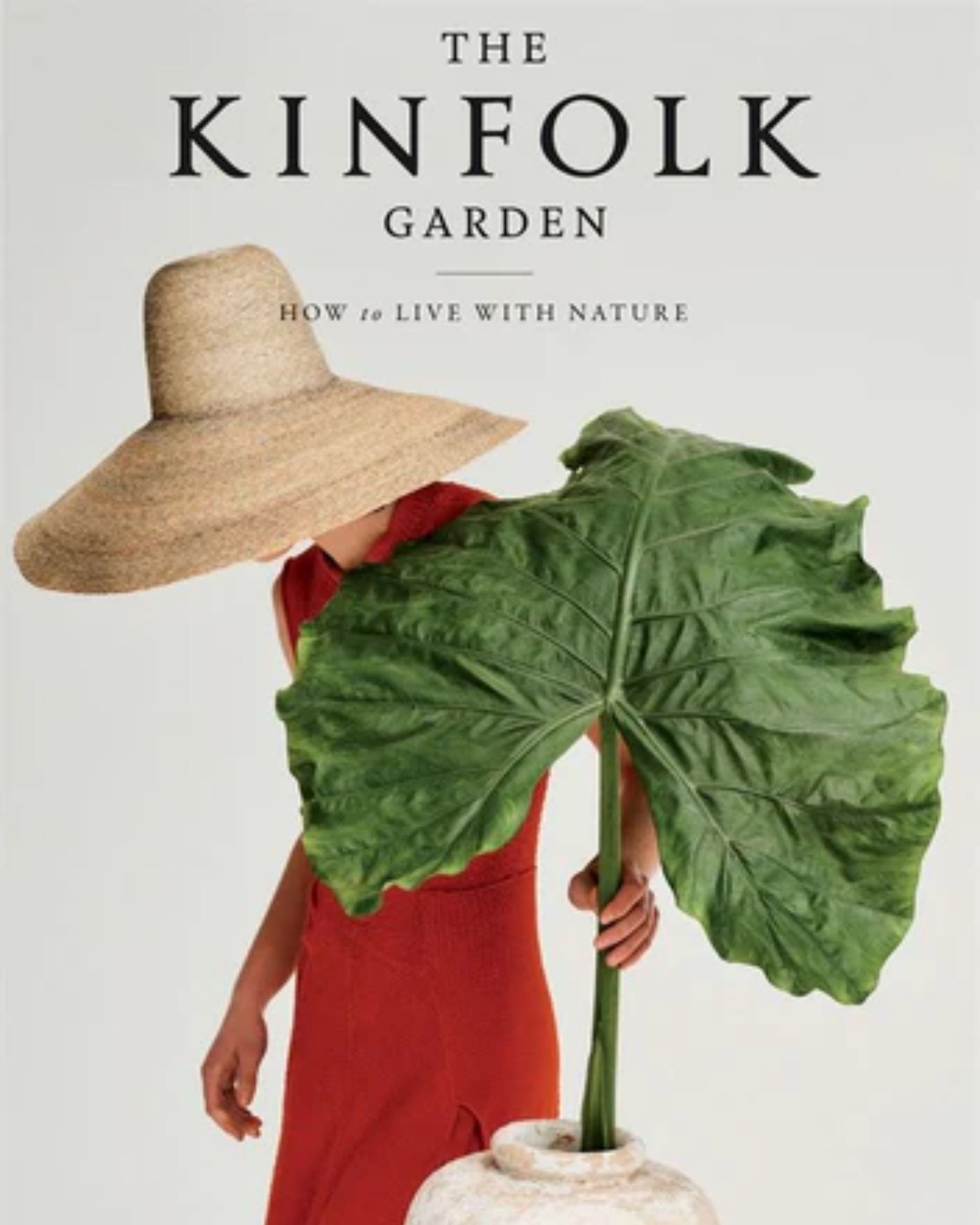 A person in a sleeveless red dress and wide-brimmed hat stands holding a large green leaf from a tall vase, taking cues from plant-filled homes. Text above reads THE KINFOLK GARDEN by Bookreps: How to Live with Nature.