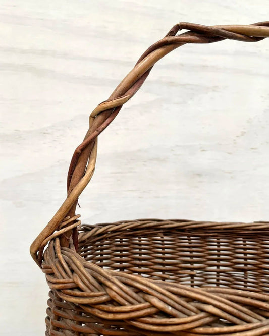 Close-up of the VINTAGE WICKER MARKET BASKET by All Things French, showcasing its handwoven natural wicker design with a twisted handle. The basket exudes rustic charm, with the handle featuring elegantly intertwined vines. Its vintage appearance is prominently highlighted against a light, neutral background.