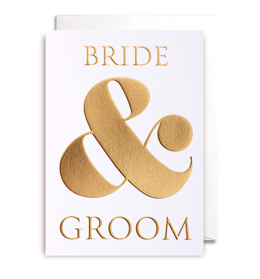 The Oxted BRIDE AND GROOM - GREETING CARD, in a pristine white design, features an elegant gold embossing of "BRIDE & GROOM" alongside a large ampersand. Slightly open, it warmly invites heartfelt wishes on its immaculate interior.