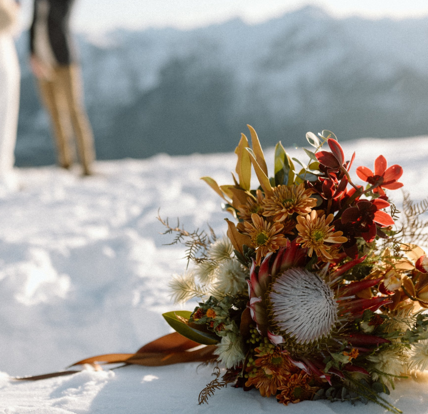 Close-up of a vibrant bridal bouquet with assorted flowers, including red, orange, and white blossoms, resting on snow. In the blurred background, two people stand on a snowy landscape with mountains visible.