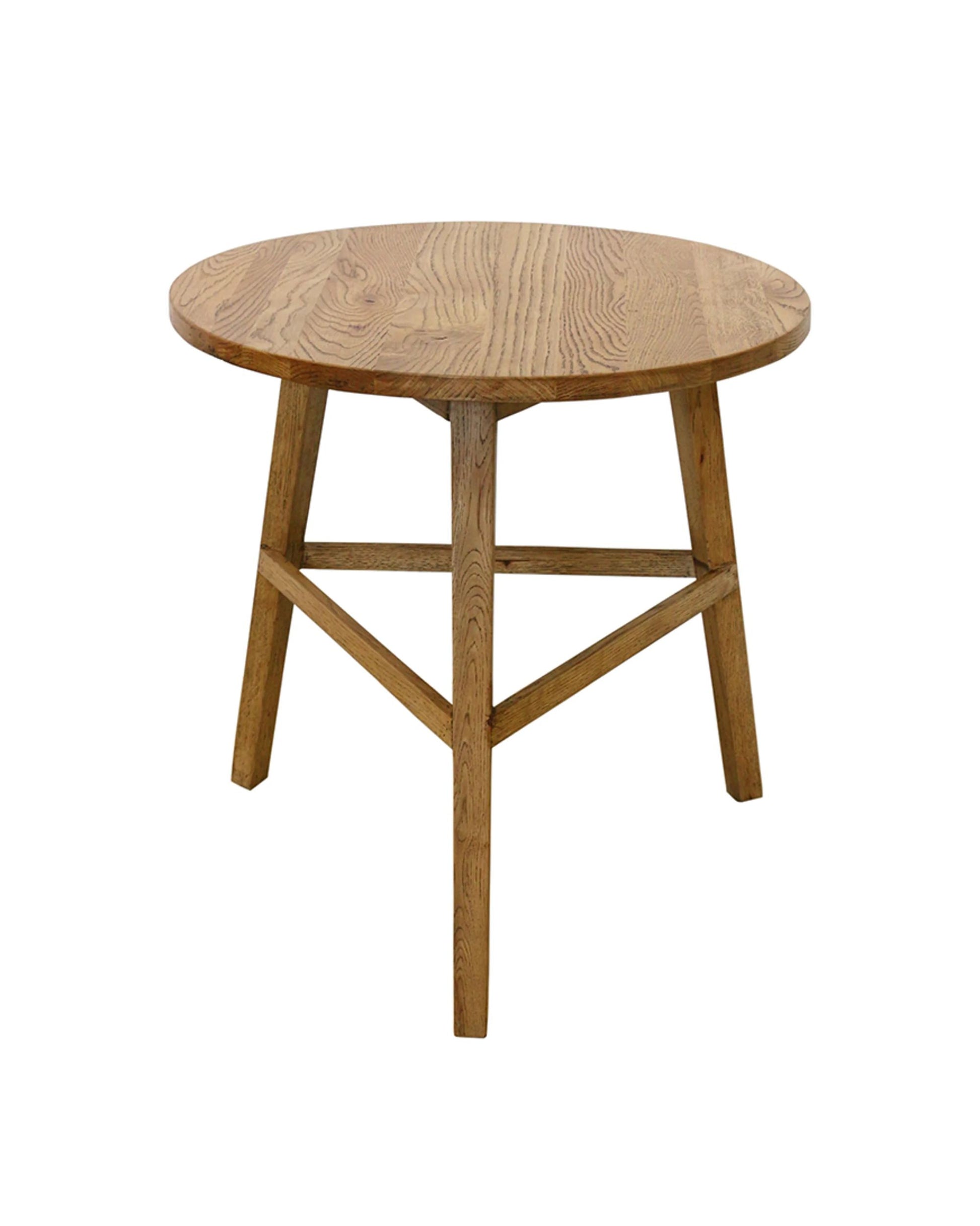 The CRICKET TABLE by CC interiors is a round solid oak table featuring a smooth surface with natural wood grain. It's supported by four sturdy legs connected by crossbars. This piece draws inspiration from traditional English cricket tables, offering a rustic design that adds elegance and suits various interior settings.