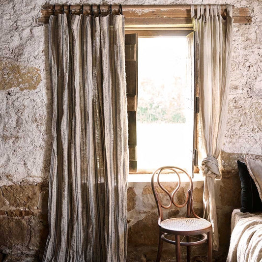 A rustic room with stone walls features an open window, adorned with Eadie's weathered Jakob Linen Curtains in a natural and charcoal stripe. A wooden chair with a circular backrest and woven seat sits in front of the window, and a dark cushion rests on the edge of a bed.