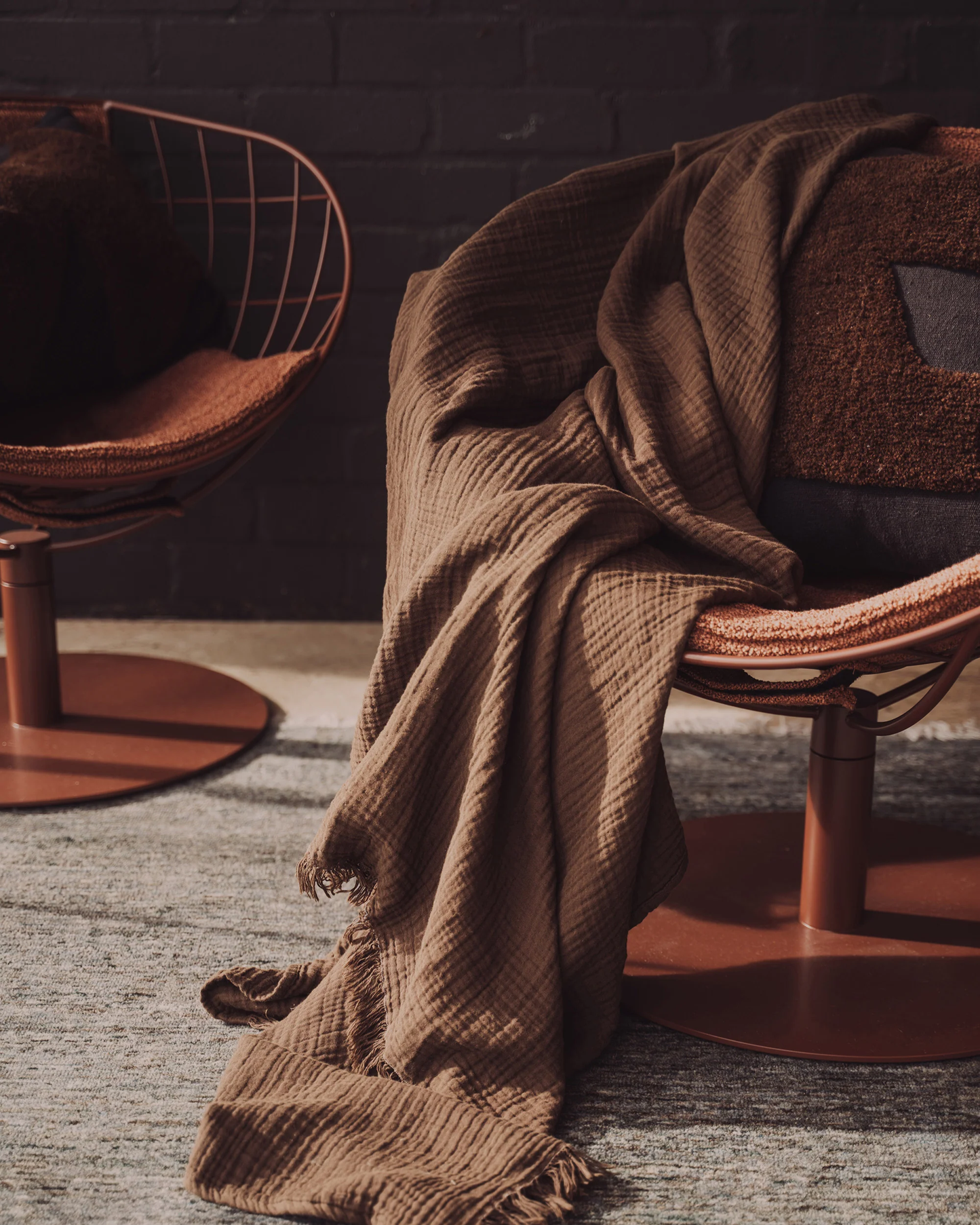 In a welcoming and stylish setup, two contemporary round chairs in a warm brown tone are elegantly adorned with the ENES COTTON THROW - CHOCOLATE by Saarde. They are positioned on a soft, light-colored rug against a dark brick wall, enhancing the cozy ambiance.