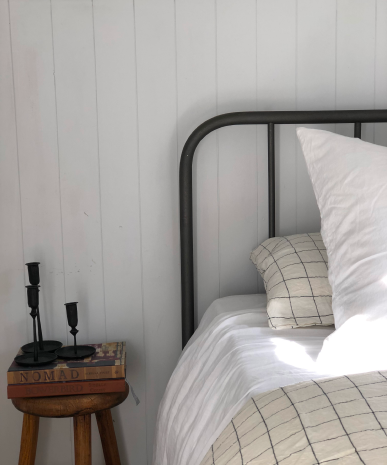 A cozy bedroom scene with a metal bed frame and white bedding featuring a black grid pattern. A wooden stool beside the bed holds two stacked books and three black candle holders, set against a white paneled wall.