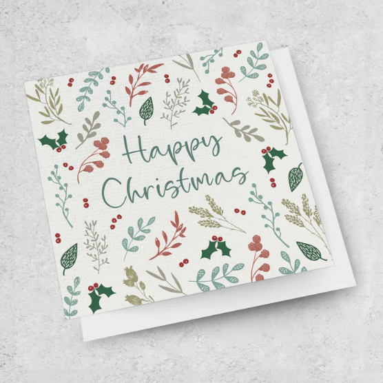 An Ink Bomb greeting card titled "HAPPY CHRISTMAS" showcases a festive design with the words "Happy Christmas" elegantly written in green script. The background is adorned with a pattern of holly leaves, berries, and various botanical elements in red, green, and beige tones.