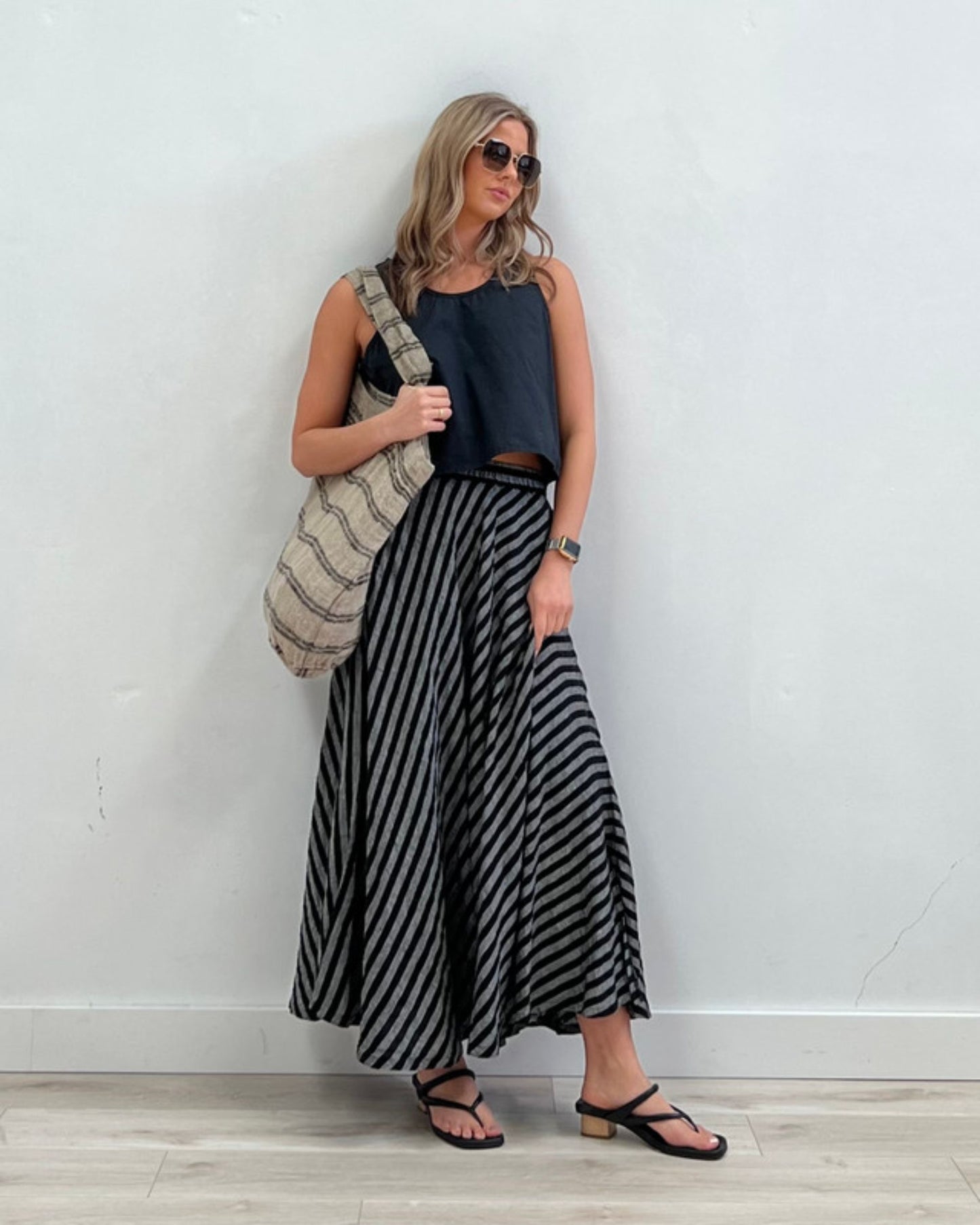 A woman in sunglasses stands against a white wall, exuding effortless style. She wears a black sleeveless top and a long, flowing skirt with black and white stripes. Carrying the Rustic Linen ANGASTON BLACK STRIPE LINE TOTE BAG, she completes the look with black sandals. Her hair is loose, one hand in her pocket.