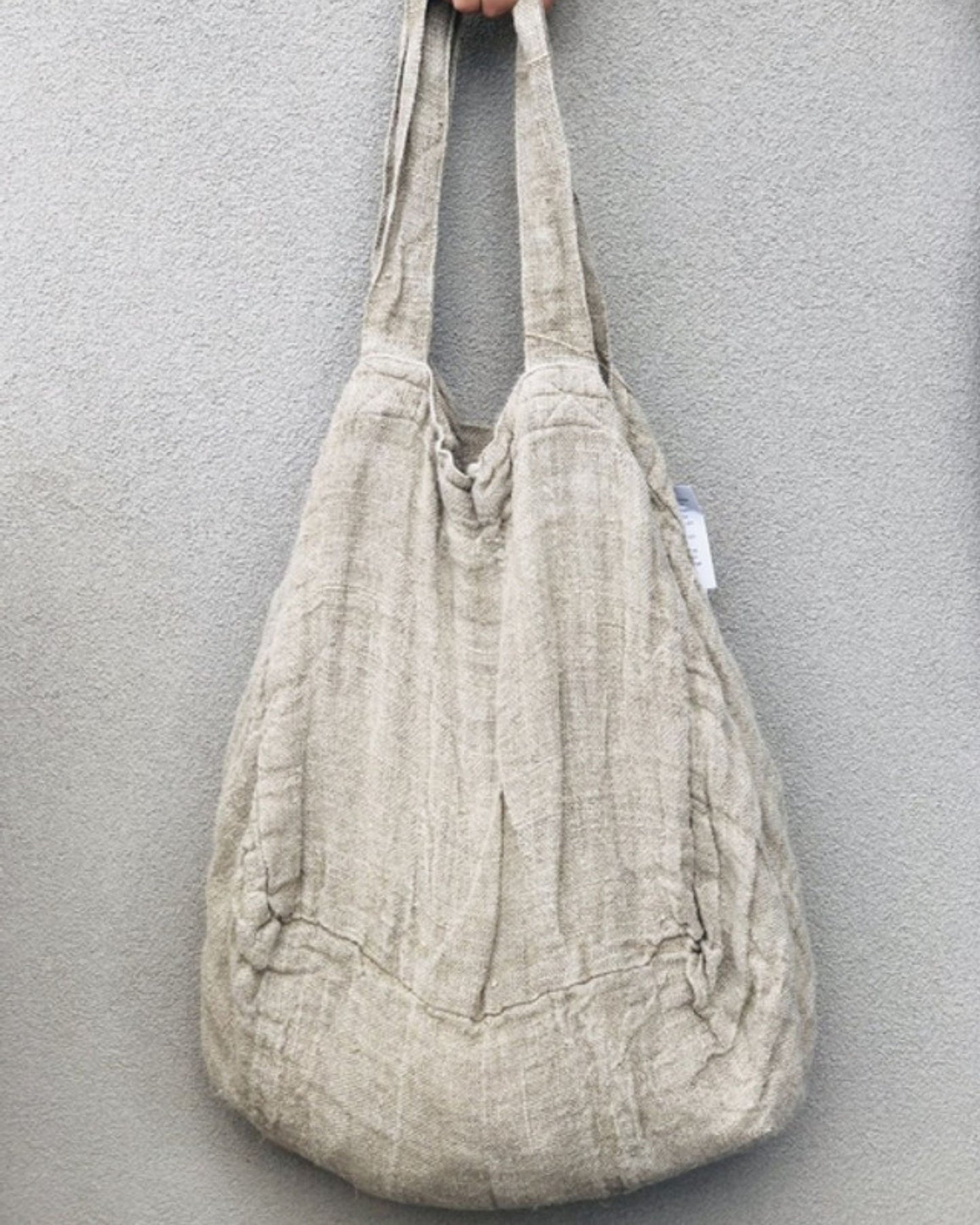 A hand holds the ANGASTON ROUNDED LARGE TOTE BAG - NATURAL by Rustic Linen, made from 100% pure linen and featuring wide handles, set against a plain grey background.