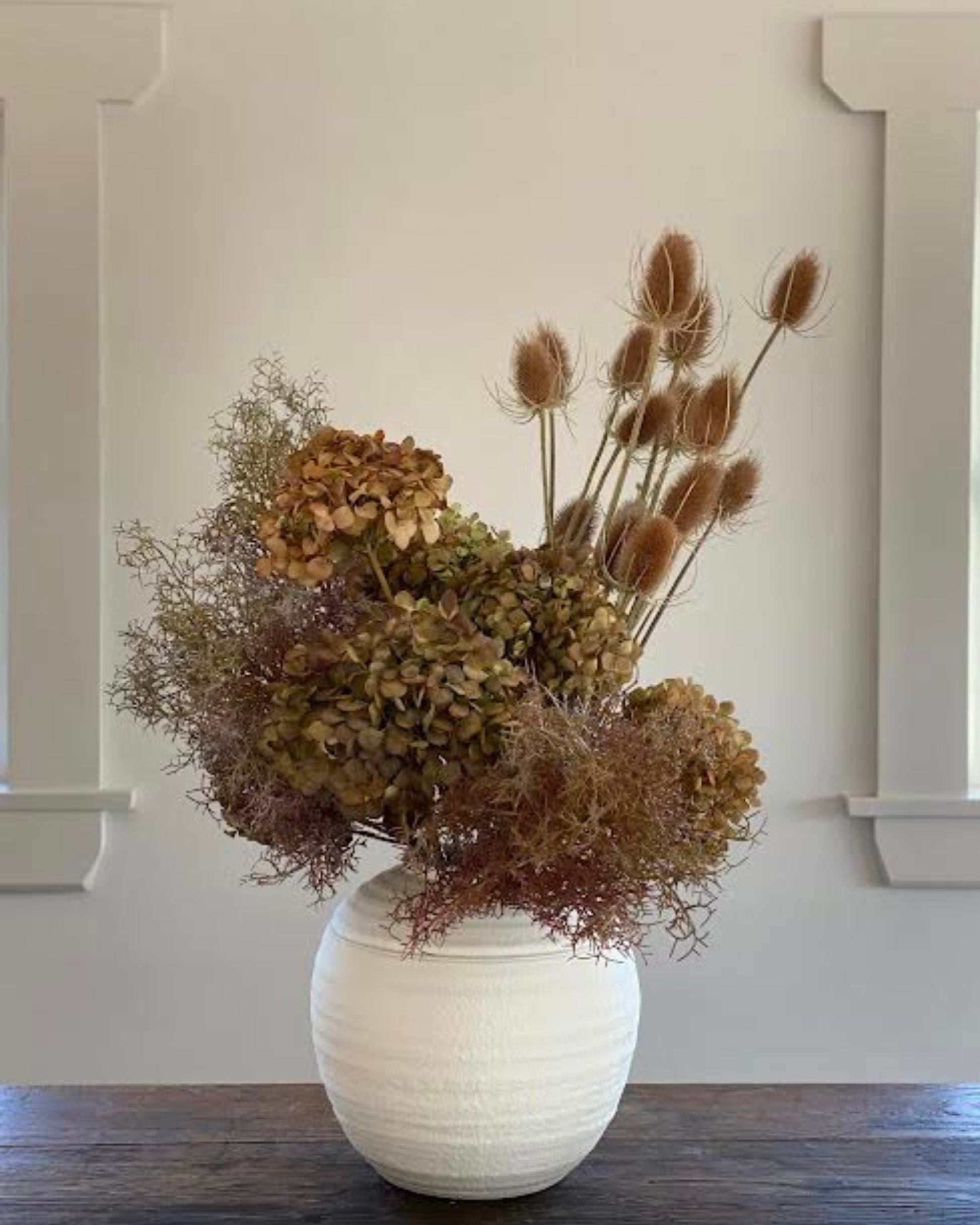 A bespoke dried flower arrangement from The Workroom NZ, featuring brown hydrangeas and thistle-like plants, is elegantly displayed in a white textured vase on a wooden surface against a light-colored wall with decorative moldings.