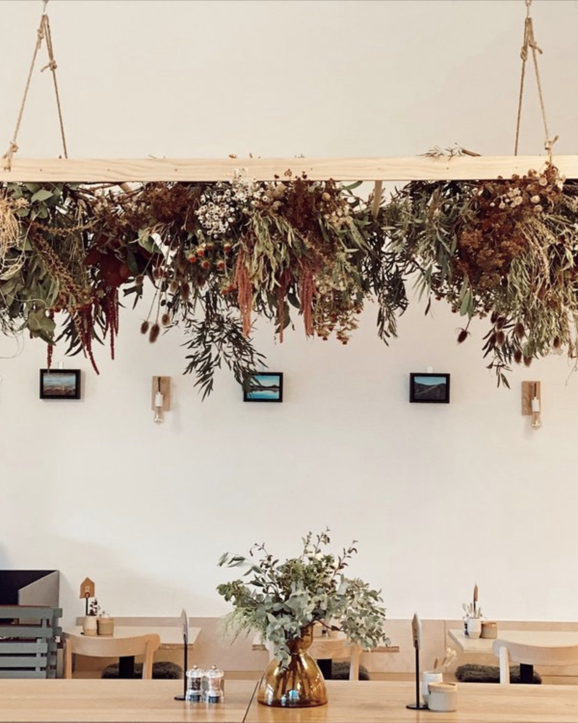 A minimalist cafe interior features a wooden table topped with a vase of greenery. Above, a BESPOKE DRIED FLOWER ARRANGEMENT by The Workroom NZ is beautifully suspended from the ceiling. The white walls are decorated with small framed pictures and wall-mounted lights.