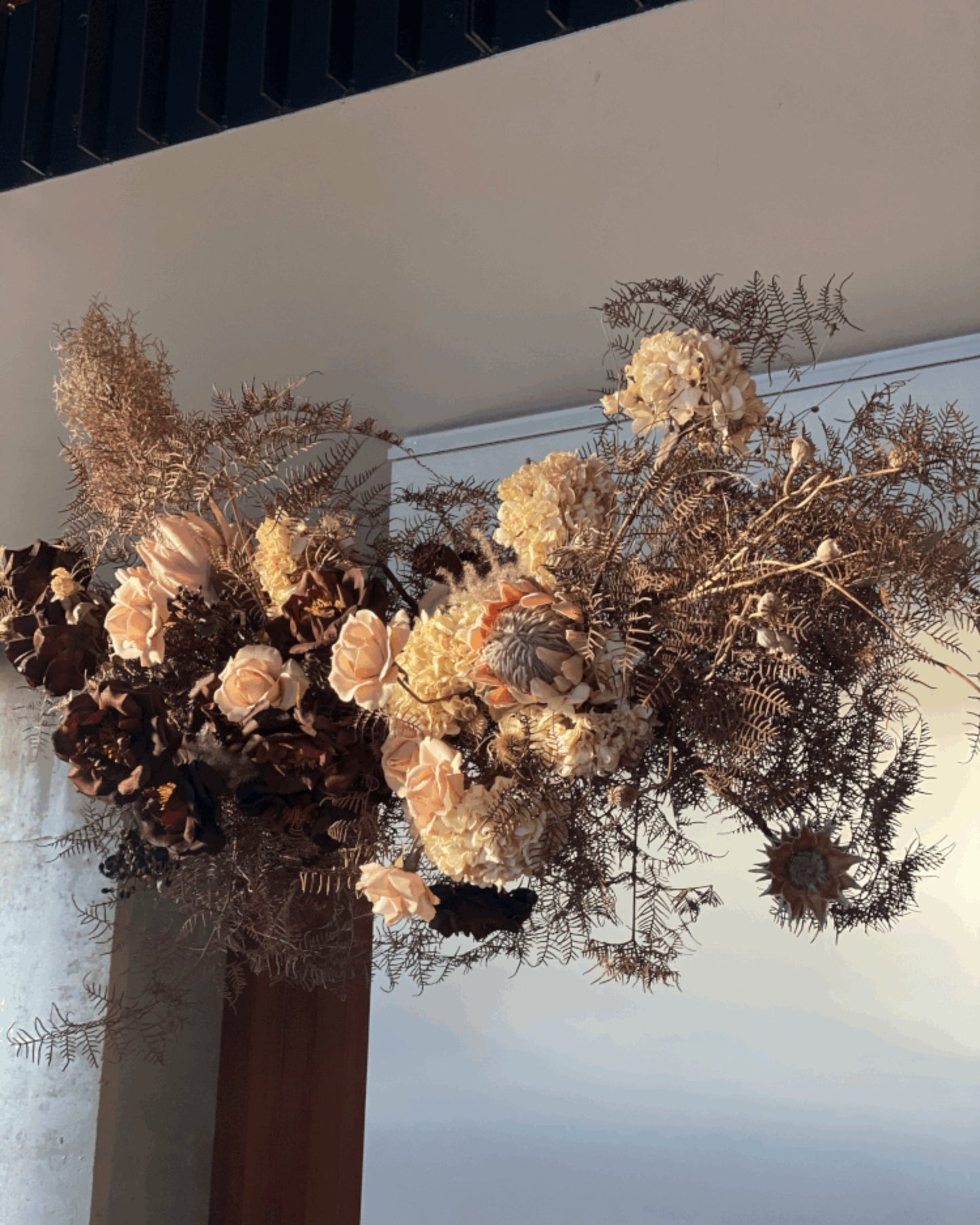 The Workroom NZ's BESPOKE DRIED FLOWER ARRANGEMENT beautifully features dried florals and ferns in earthy hues such as light peach, brown, and beige, elegantly set against a neutral backdrop to emphasize textures and natural beauty.