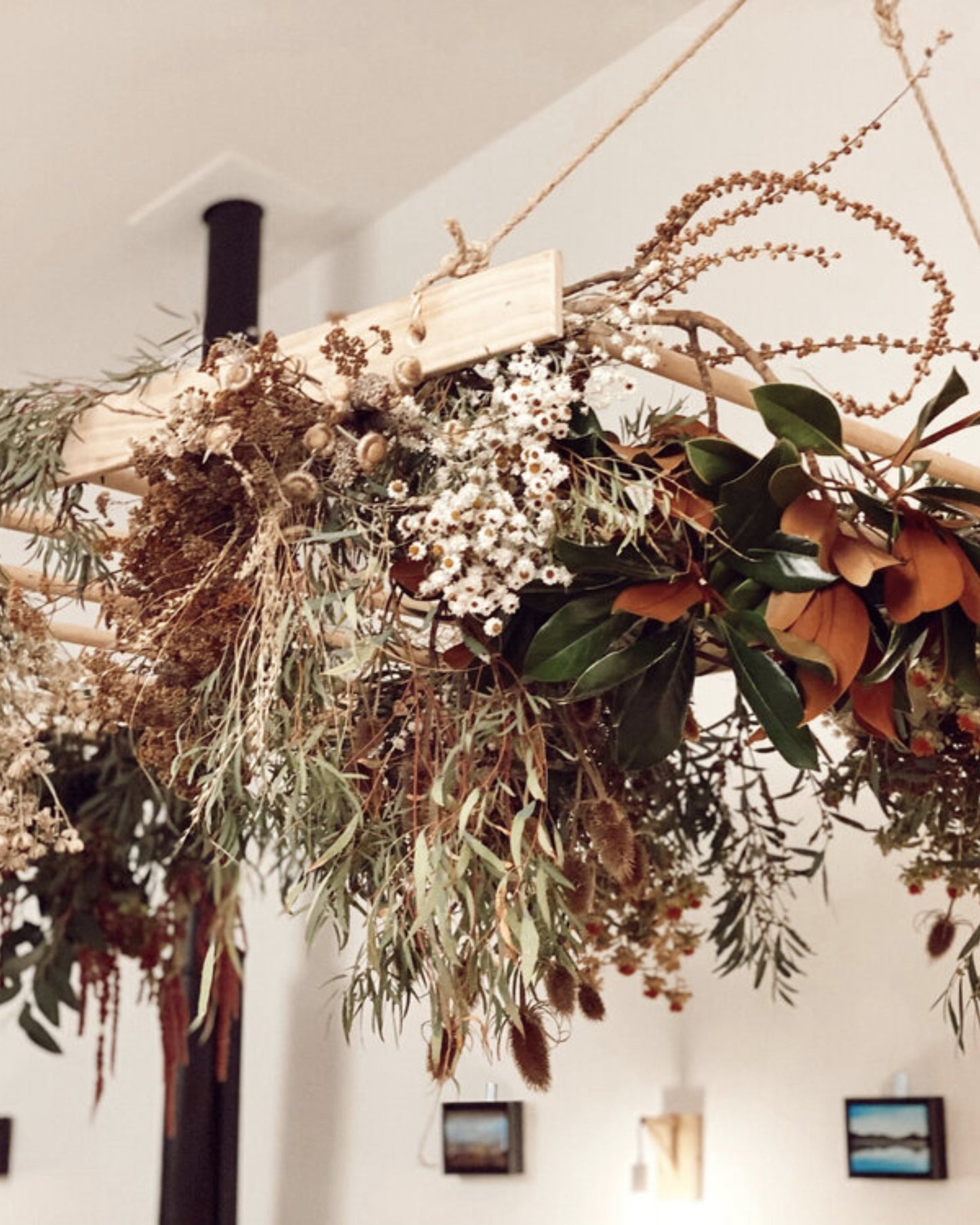 A custom creation by The Workroom NZ, the BESPOKE DRIED FLOWER ARRANGEMENT elegantly displays dried florals and lush green foliage, artistically suspended from a wooden frame. The setting features a light-colored wall adorned with small framed pictures, adding an extra touch of charm.