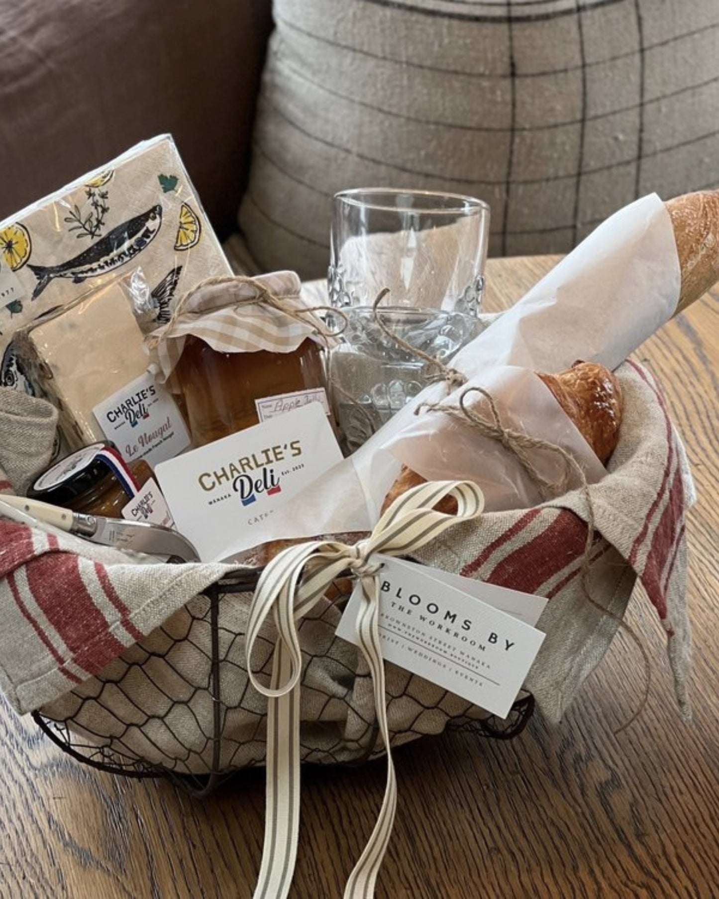 At The Workroom, a FRENCH LOVERS PICNIC GIFT BASKET filled with a baguette, croissant, artisanal homemade fruit spread, and cheese awaits you. This rustic delight is elegantly lined with cream and red cloth and features a charming Bloom label, all set on a wooden table for your enjoyment.